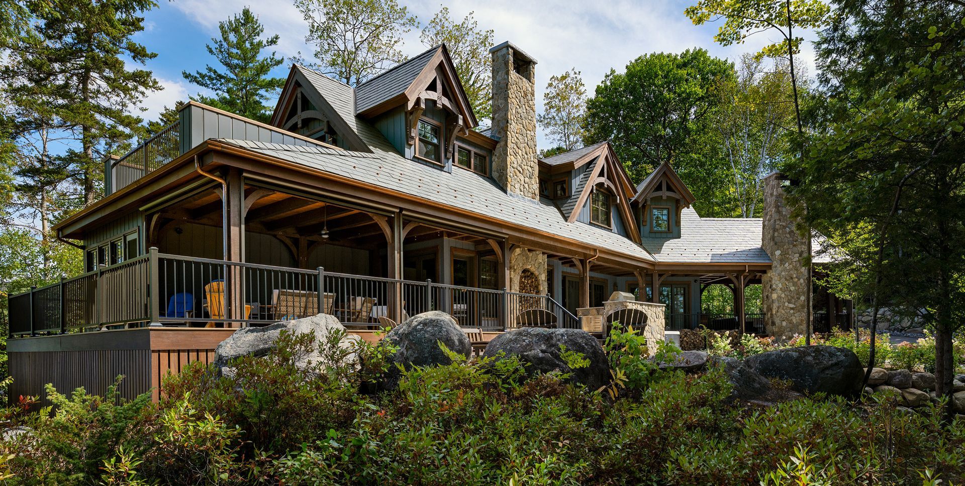 A large, rustic home with a stone chimney, wrap-around porch, and metal roof surrounded by lush trees and greenery.
