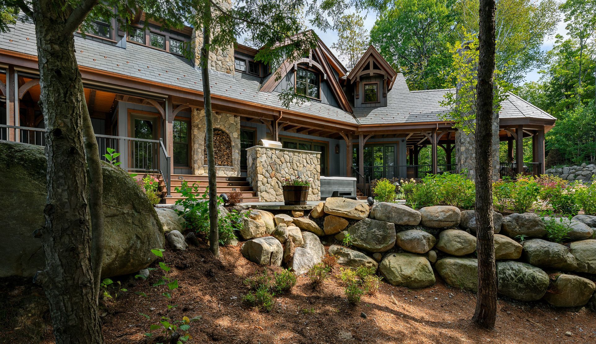 A large, rustic house with a stone foundation and slate roof, set in a wooded area with a stone wall in the foreground.