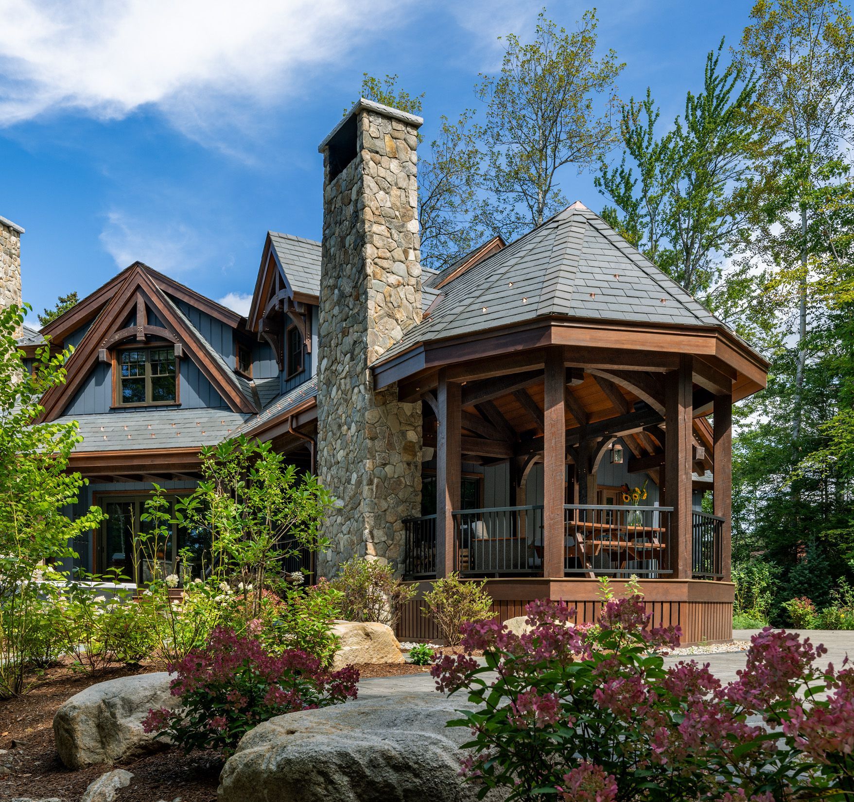 A stone and wood luxury house with a prominent chimney and a covered, octagonal porch surrounded by landscaped gardens.