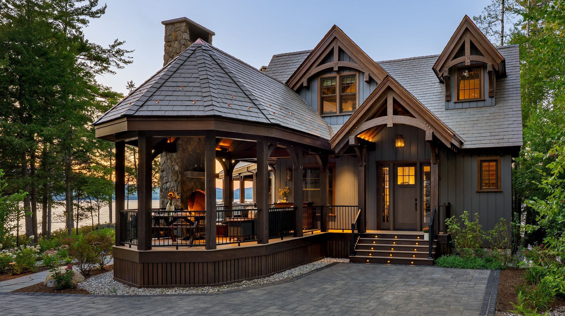 A cozy cottage with a stone chimney, wrap-around porch with a fireplace, and steep shingled roof, set in a wooded area.