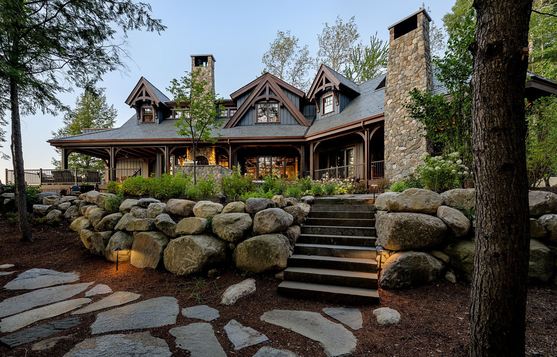 A rustic, multi-gabled wooden home with stone chimneys overlooks a stone retaining wall, patio, and steps in a forest.