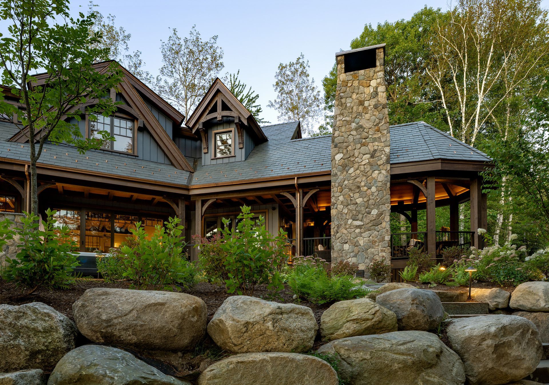 A rustic cabin with a stone chimney and covered patio, situated behind a large stone retaining wall and lush landscaping.