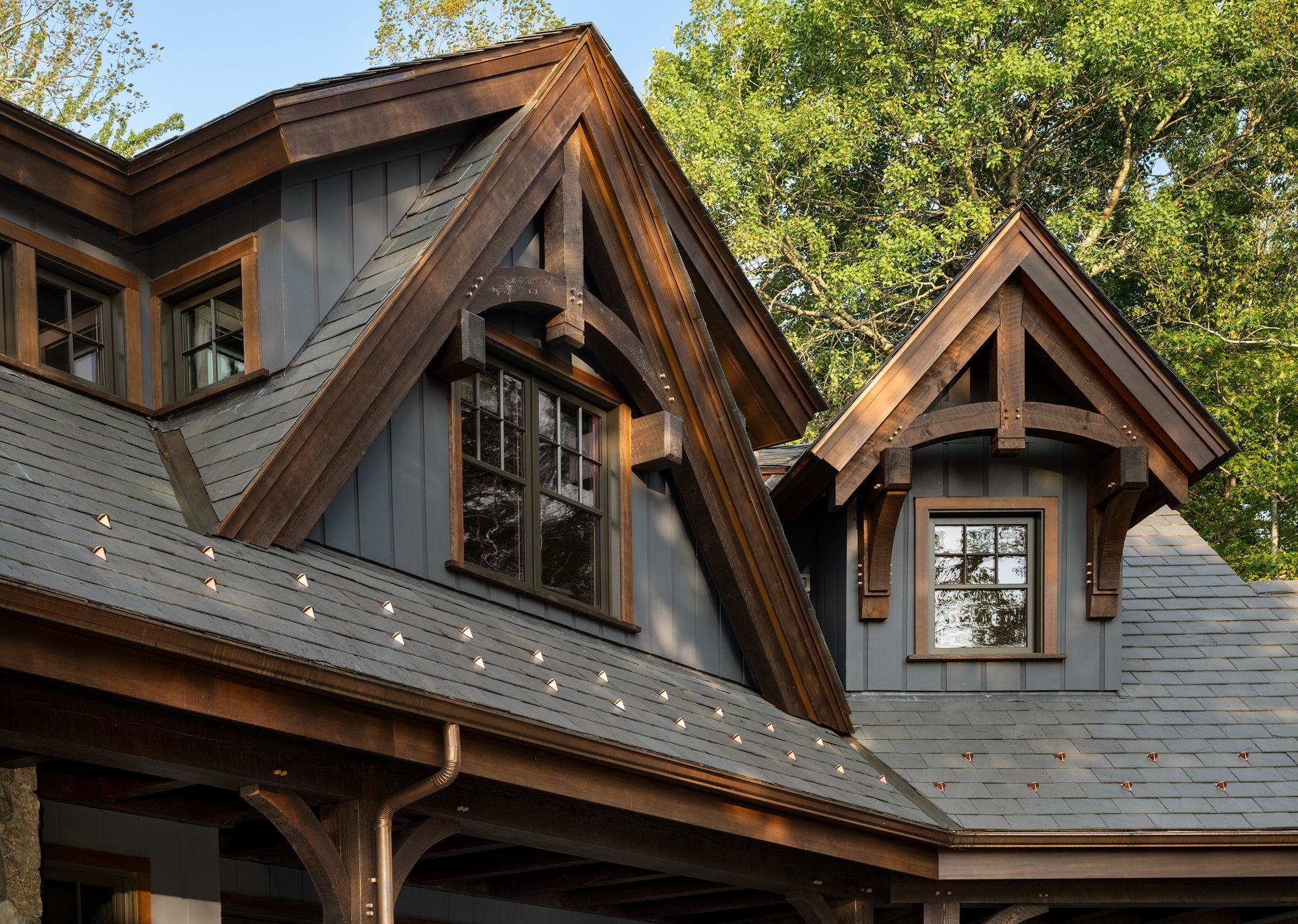 An architectural view of a house exterior featuring dark wood timber framing, gray siding, and gabled roofs among trees.