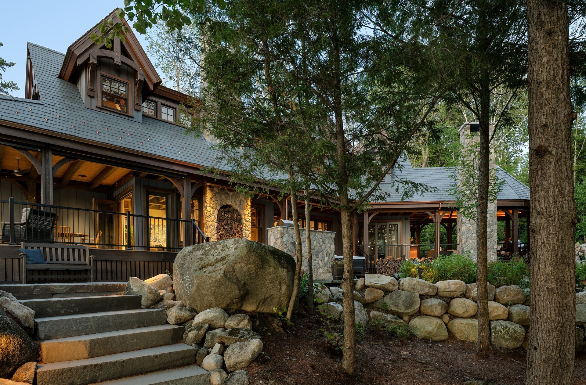 A rustic home with a stone chimney and wraparound porch, featuring a tiered stone staircase and a stone wall in a forest.