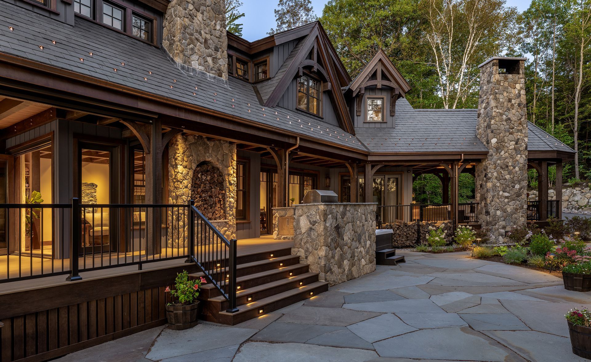 A rustic, multi-story stone and wood mountain home with a large paved patio, stairs, and a wooden porch at dusk.