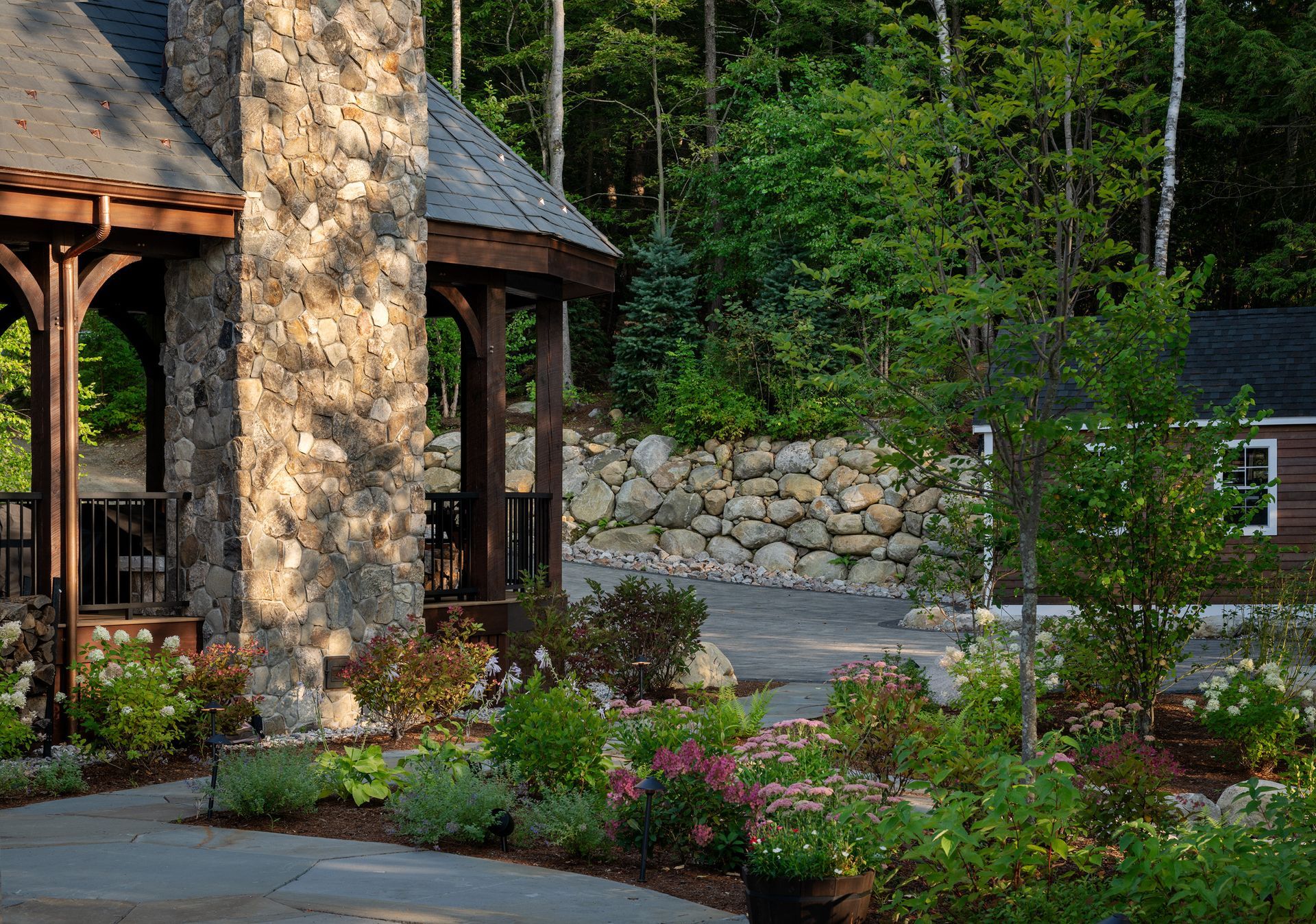 A stone gazebo with a dark roof stands beside a lush garden and a stone retaining wall in a sunlit, wooded setting.