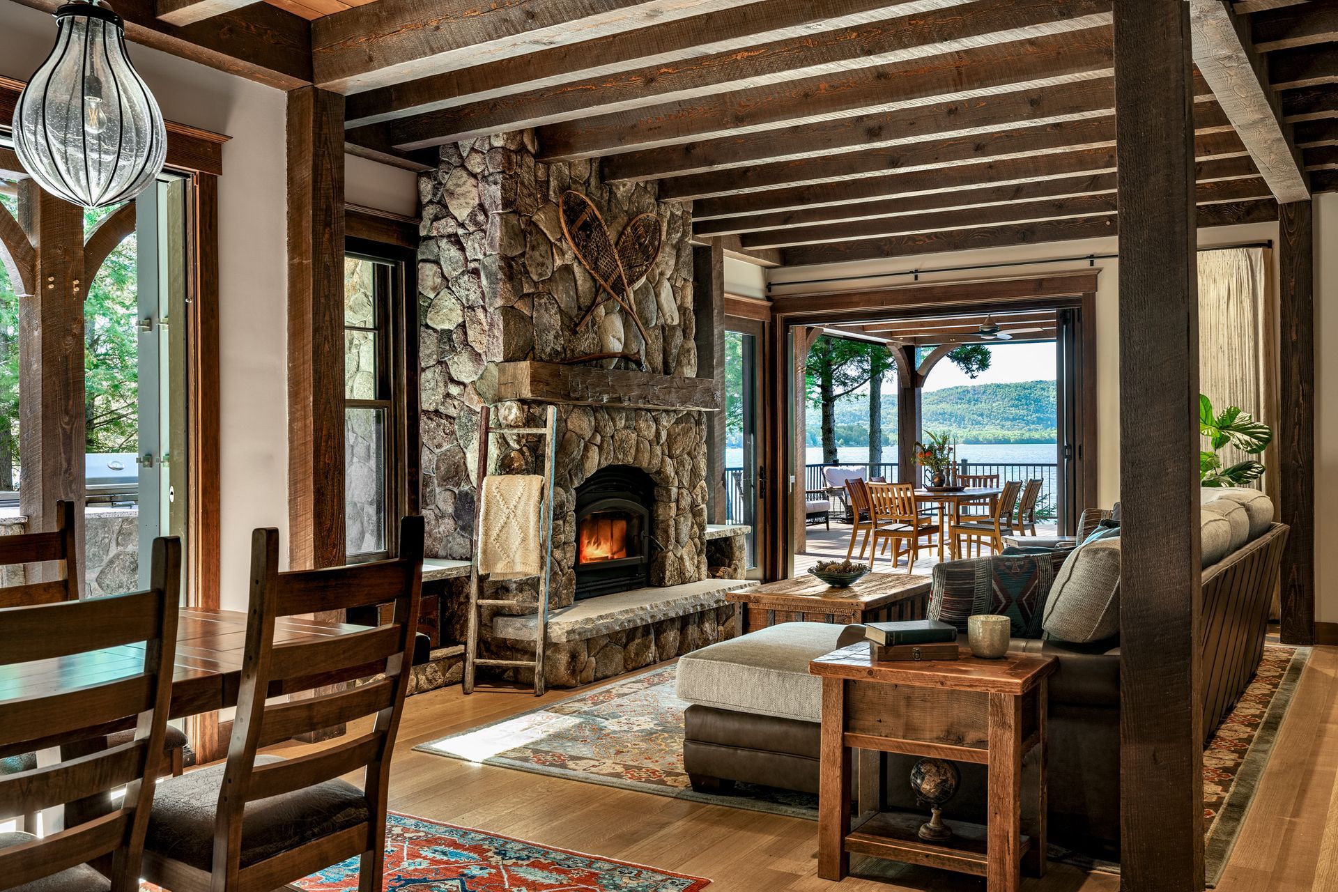 Rustic living room with exposed wood ceiling beams, a stone fireplace, and a view of a deck and lake through glass doors.