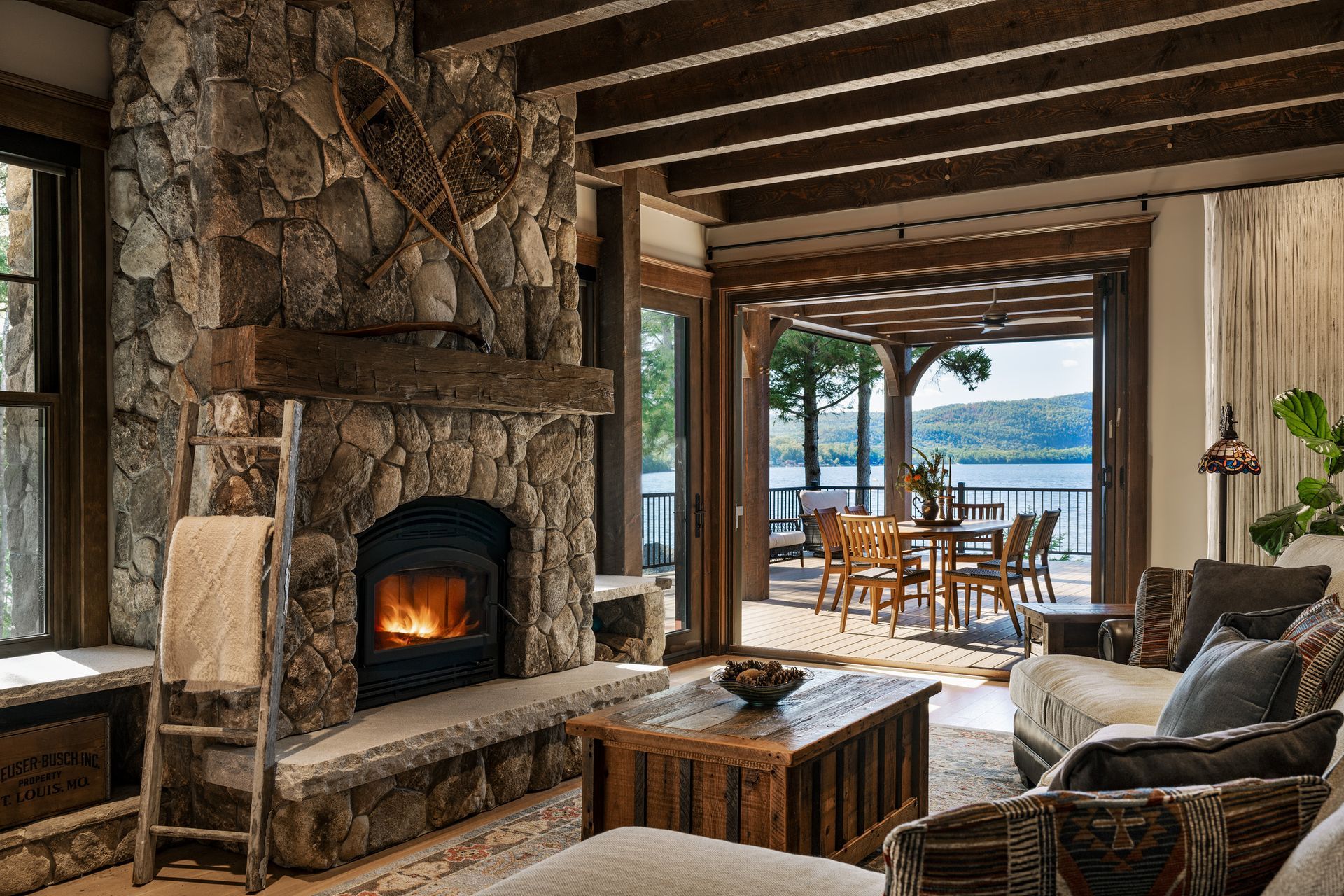 A cozy living room with a large stone fireplace, exposed ceiling beams, and a view of a lake from the back patio.
