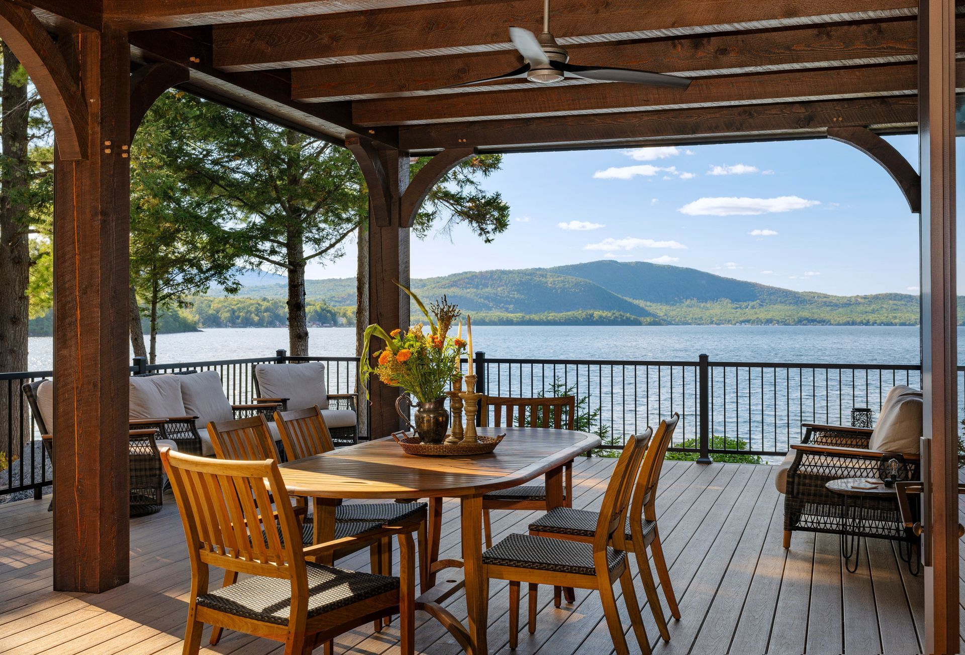 A wooden dining set on a covered deck overlooking a calm lake and distant mountains on a sunny day.