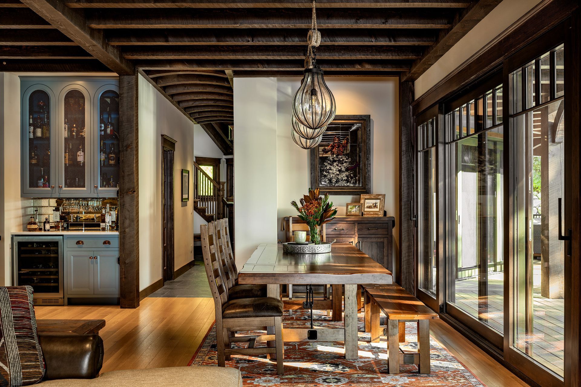 Rustic dining area with a long wooden table, patterned rug, blue cabinets, and floor-to-ceiling glass sliding doors.