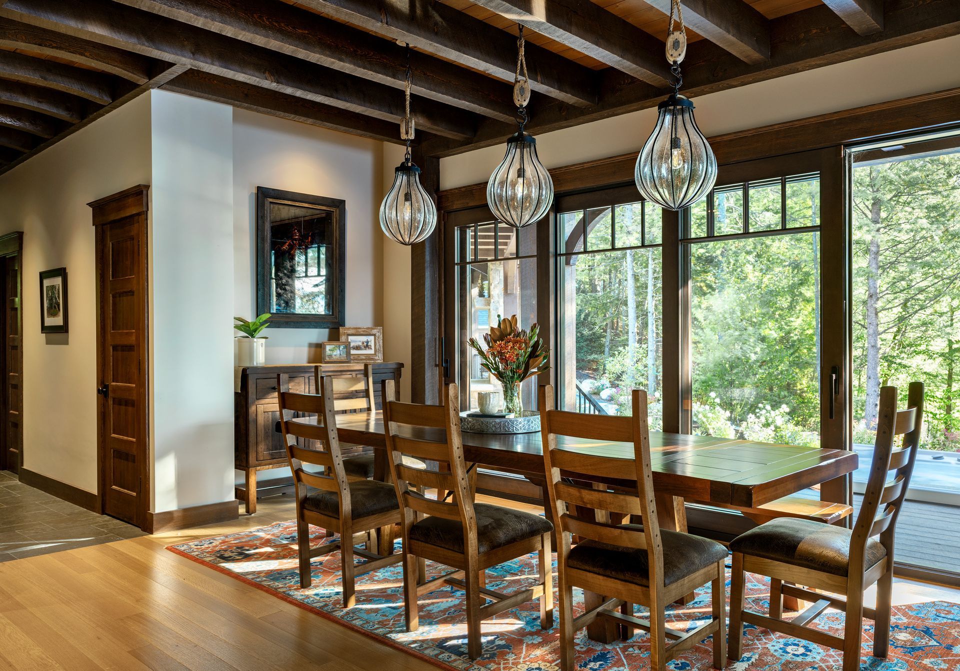 A dining room with a wooden table, six chairs, and three glass pendant lights under a beamed ceiling near large windows.