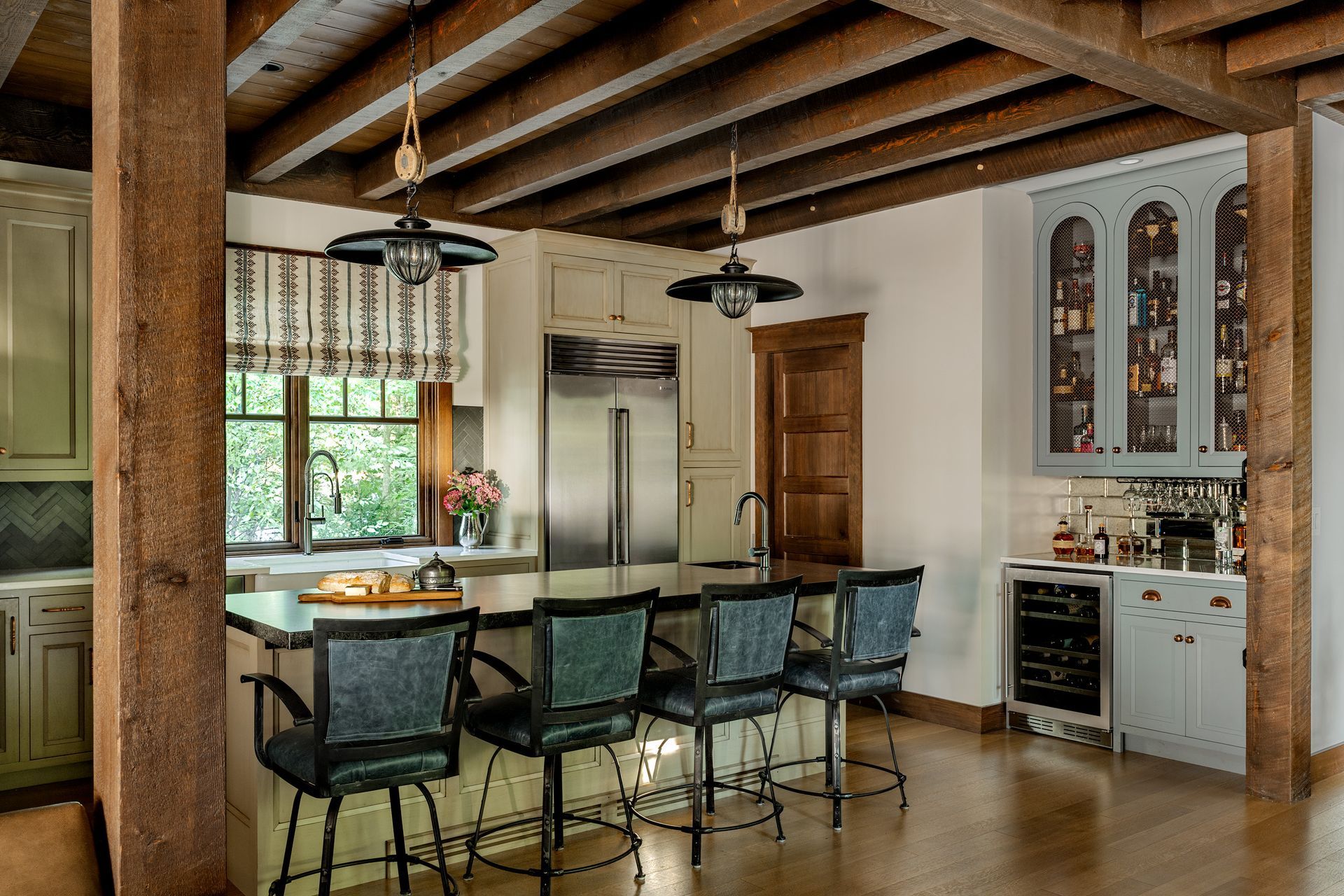 A rustic kitchen with wood ceiling beams, four bar stools at an island, and blue cabinetry with a wine cooler.