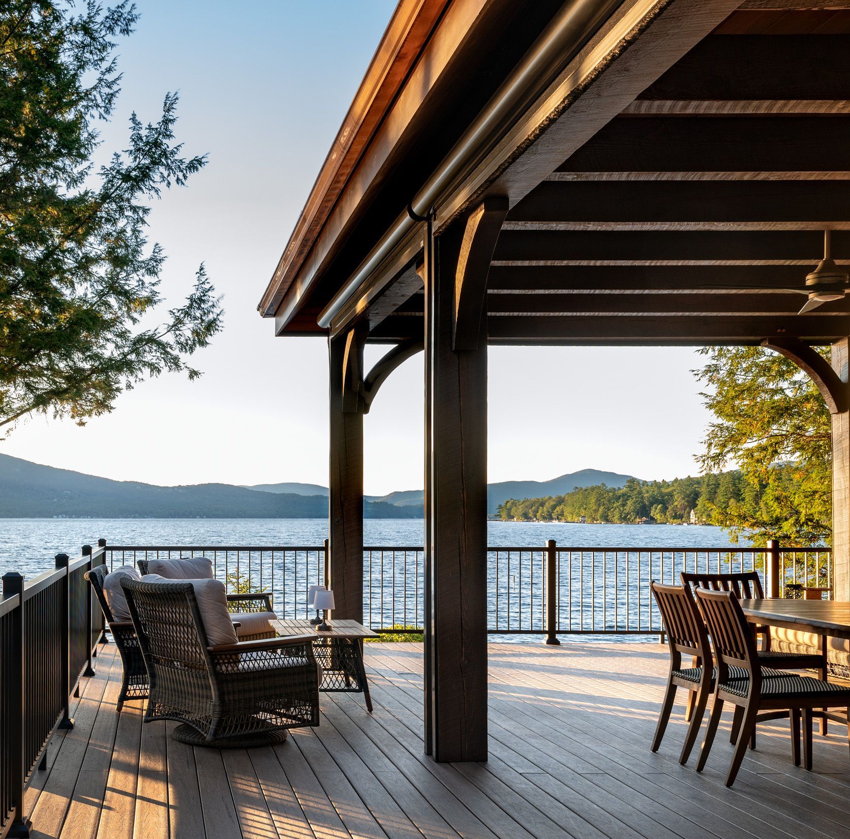 Covered deck overlooking a lake, with wicker chairs and a dining table under a dark wood structure at sunset.