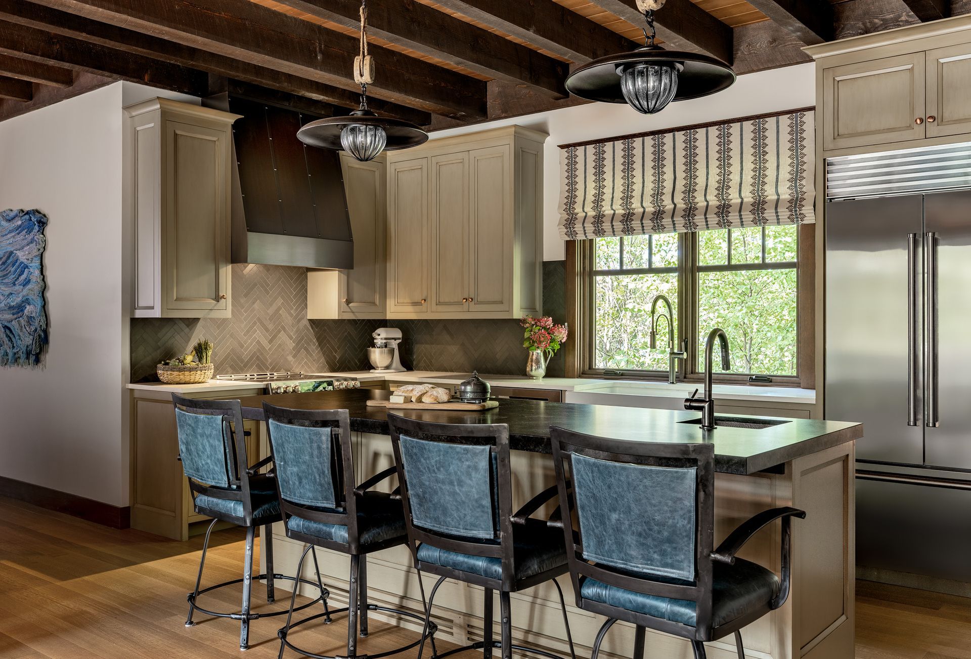 A kitchen island with four blue swivel stools sits in a modern, rustic-style kitchen with wood beams and a large fridge.