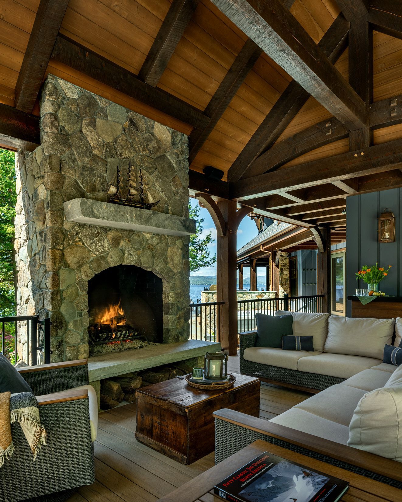 A cozy covered deck featuring a large stone fireplace, dark wood-beamed ceiling, and comfortable outdoor furniture.
