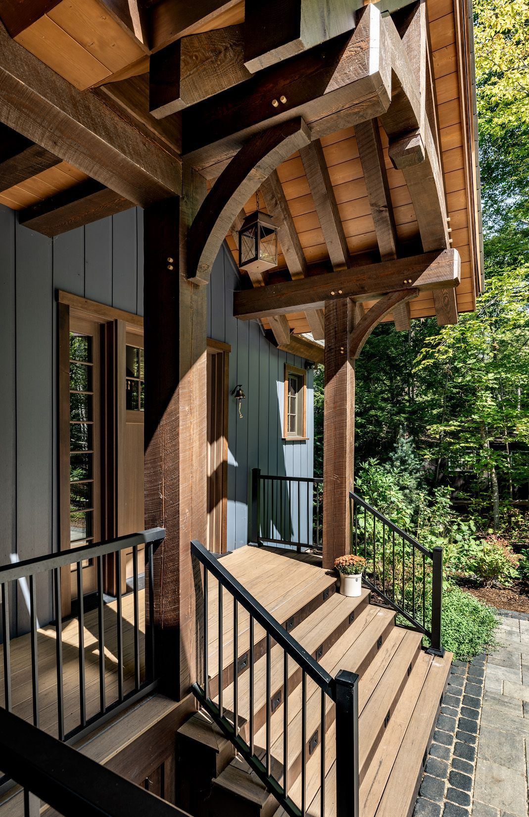 A wooden staircase with black railings leads up to a covered porch entrance on a modern gray house surrounded by trees.