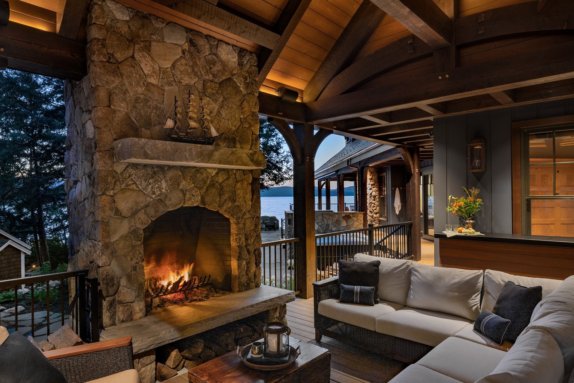A cozy outdoor covered patio featuring a large stone fireplace, wicker seating with cushions, and a waterfront view.