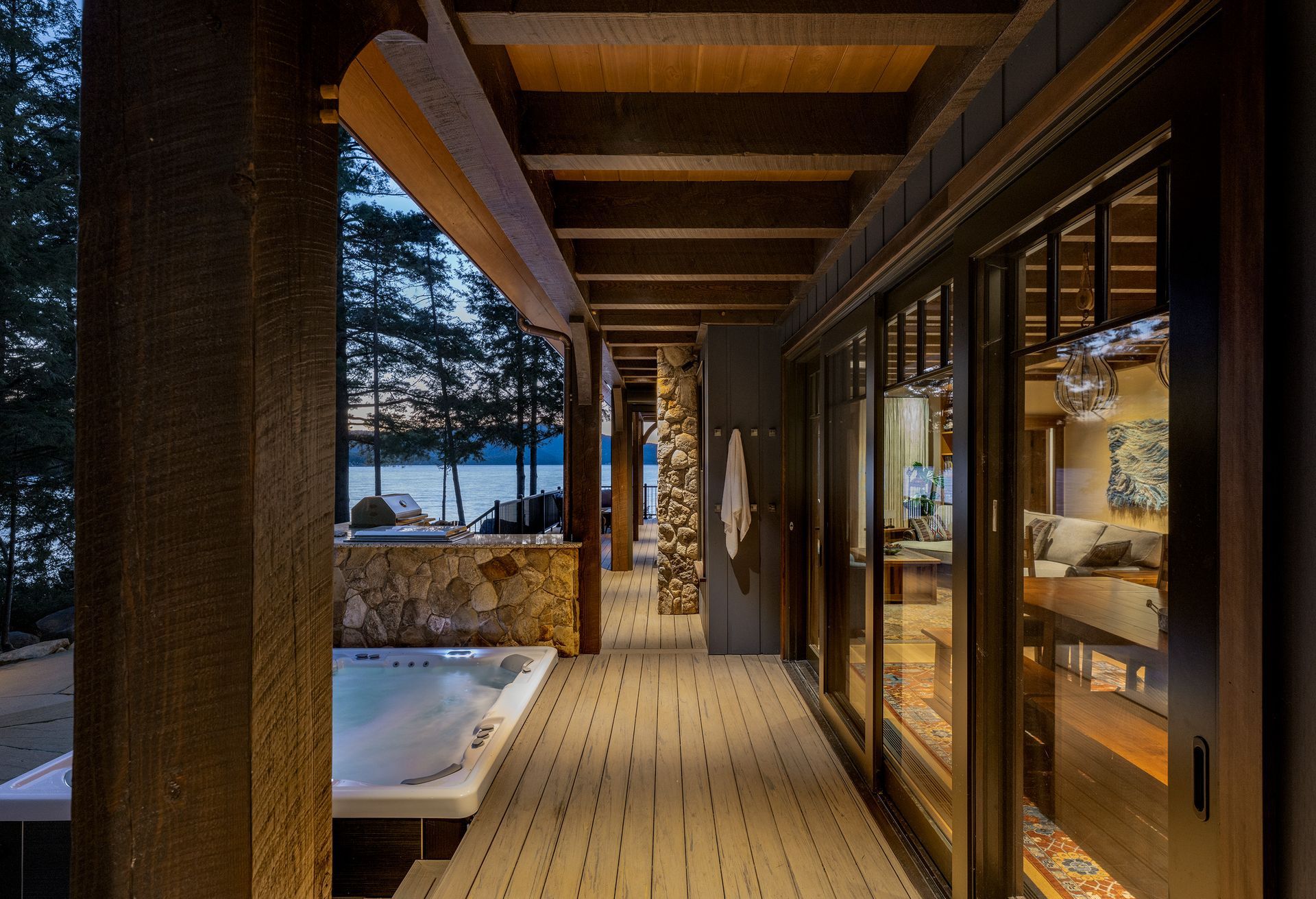 A wooden deck with a hot tub overlooks a lake at twilight, adjacent to a house with large glass doors.