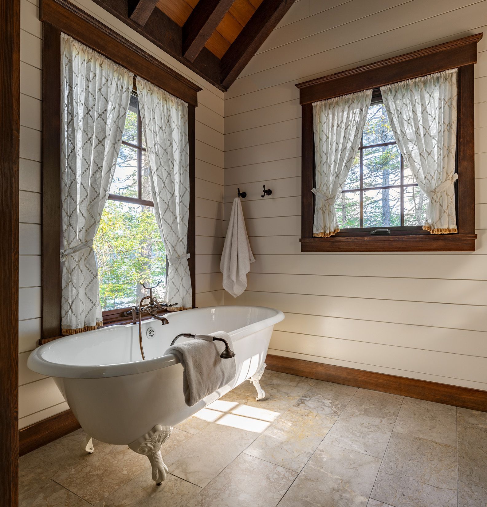 A classic clawfoot bathtub sits under two wood-trimmed windows in a rustic bathroom with white shiplap walls and tile floor.