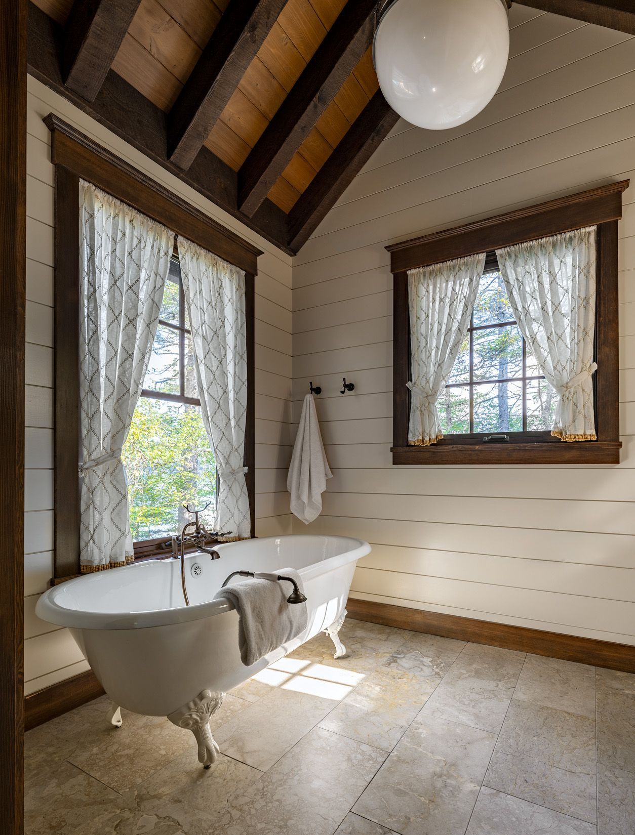 A rustic bathroom with a white claw-foot tub, vaulted wooden ceiling, paneled walls, and two windows with patterned curtains.