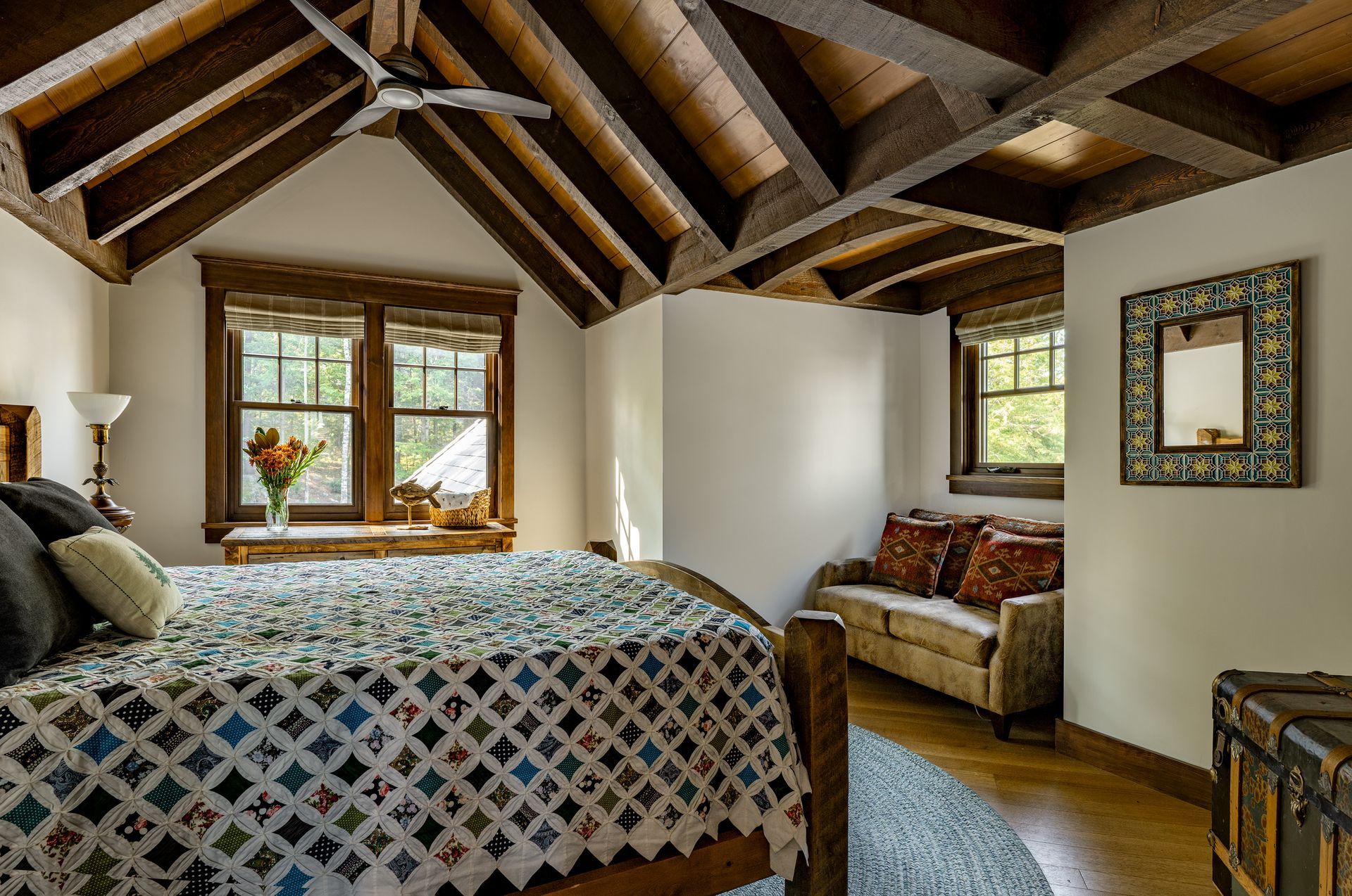 A bedroom with vaulted, beamed ceilings, a quilt-covered bed, a window with a view, and a small sofa in the corner.