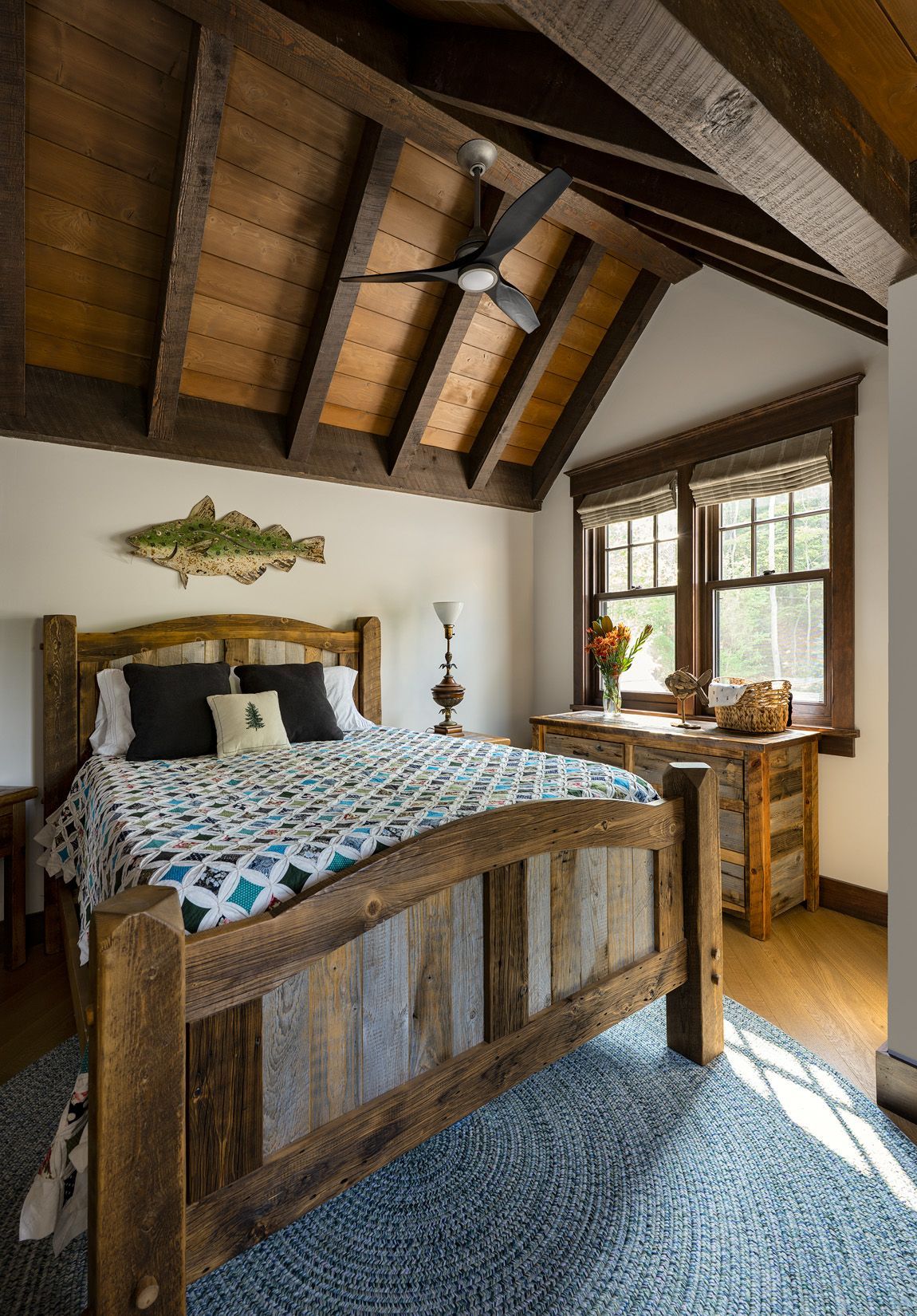 A rustic bedroom with a wooden bed, patterned quilt, exposed wood-beam ceiling, and a decorative fish on the wall.