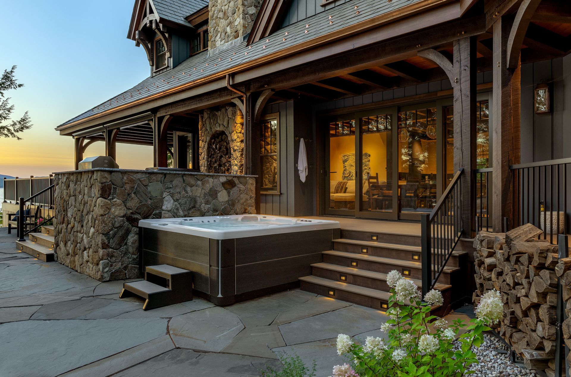 A luxury stone and wood cottage exterior featuring a hot tub on a flagstone patio at twilight.