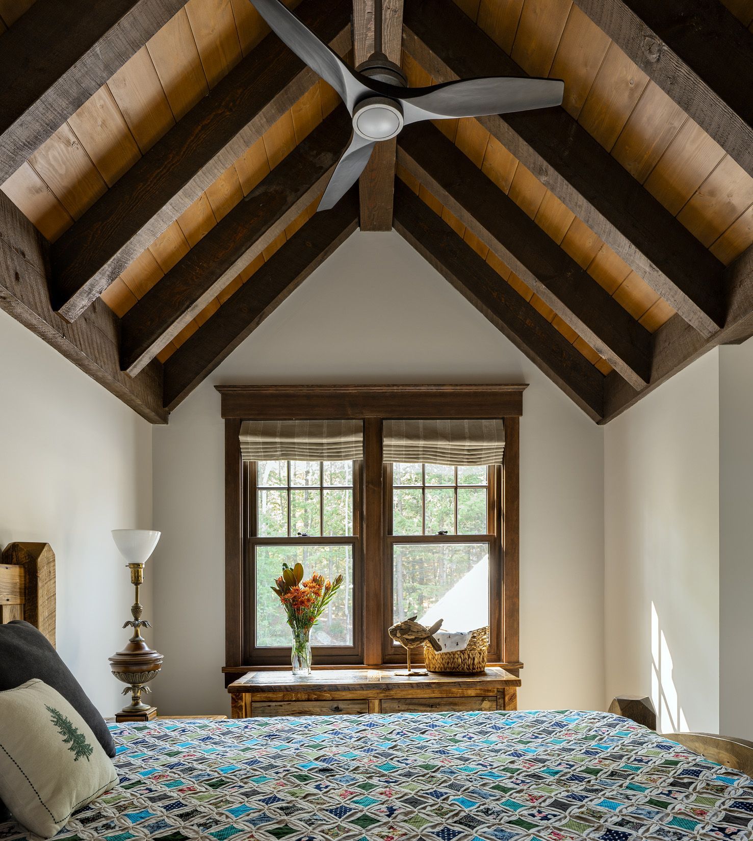 A rustic bedroom with a quilt-covered bed, a window with flowers, and exposed wooden beams on a high, vaulted ceiling.