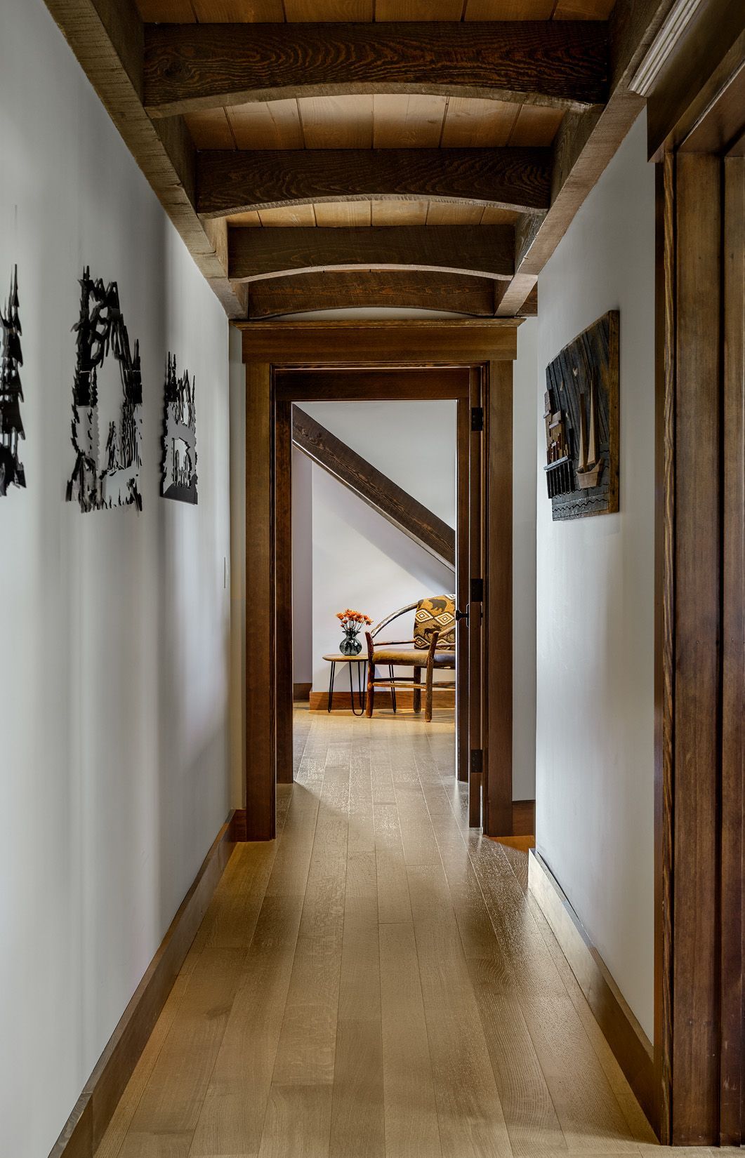 A wood-floored hallway with exposed ceiling beams leads to another room containing furniture and a staircase.
