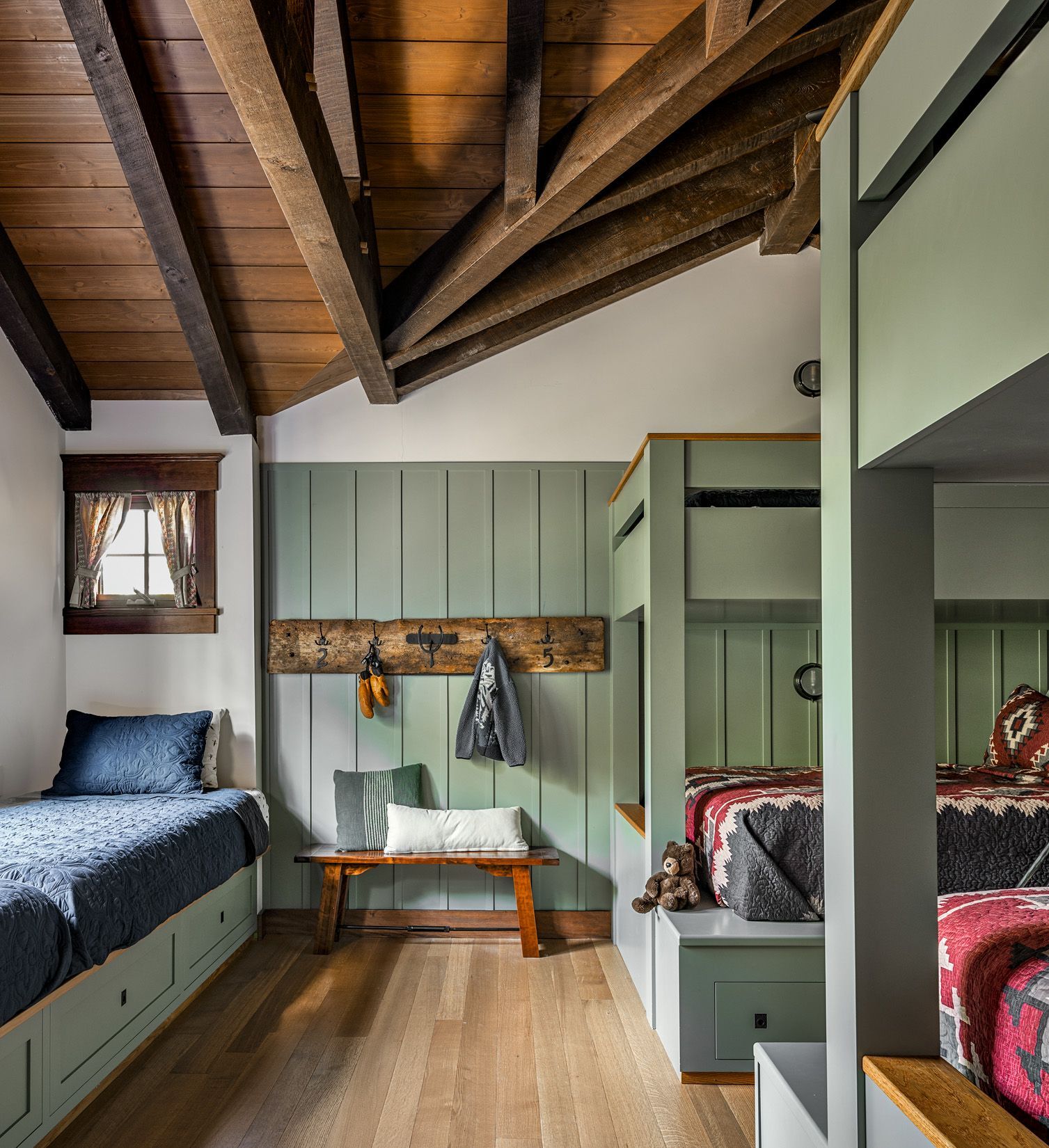A rustic bedroom with wood ceiling beams, a daybed, a wooden bench against sage green walls, and built-in bunk beds.