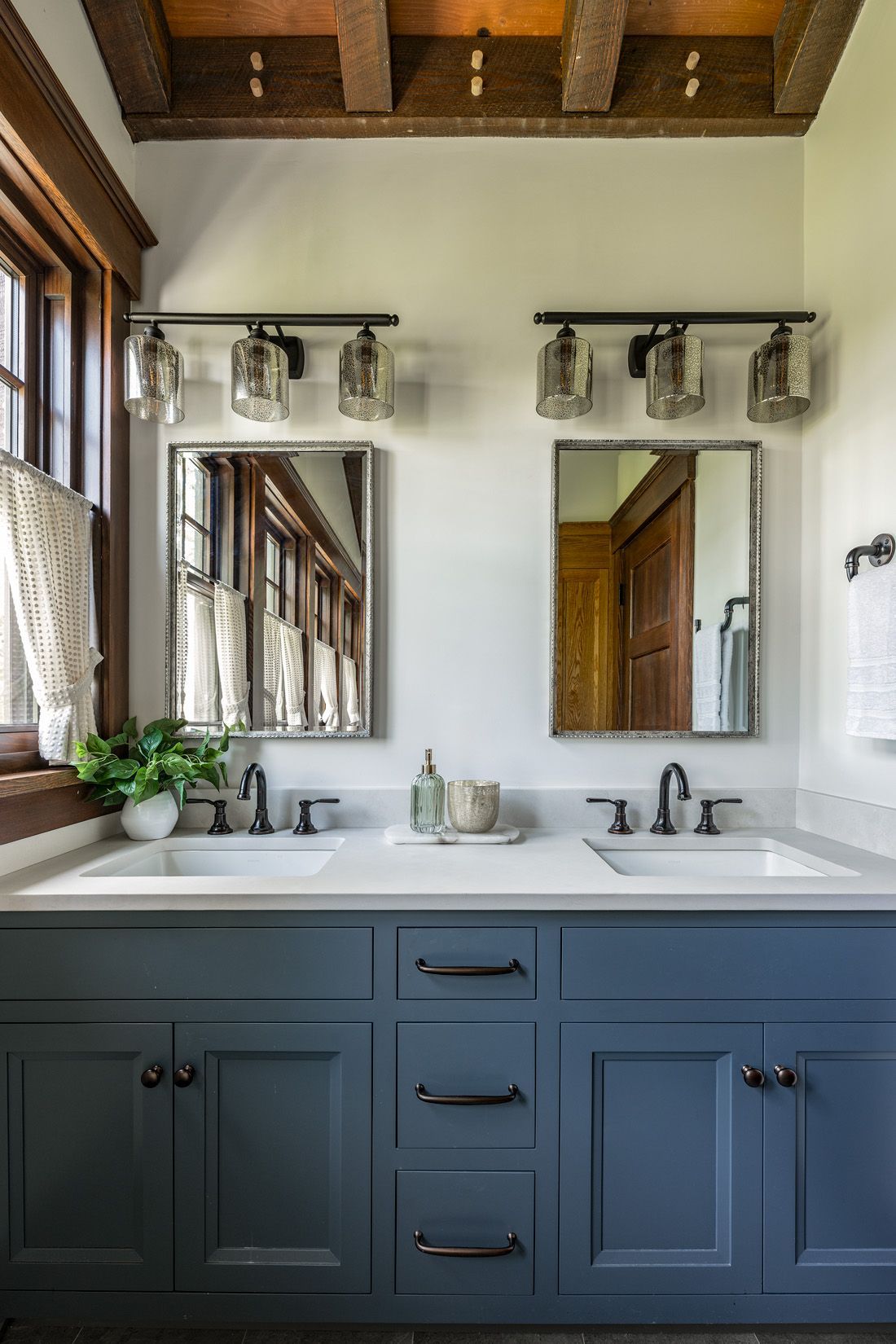 A double-sink bathroom vanity with blue cabinets, two mirrors, and industrial-style lighting under a rustic wood ceiling.