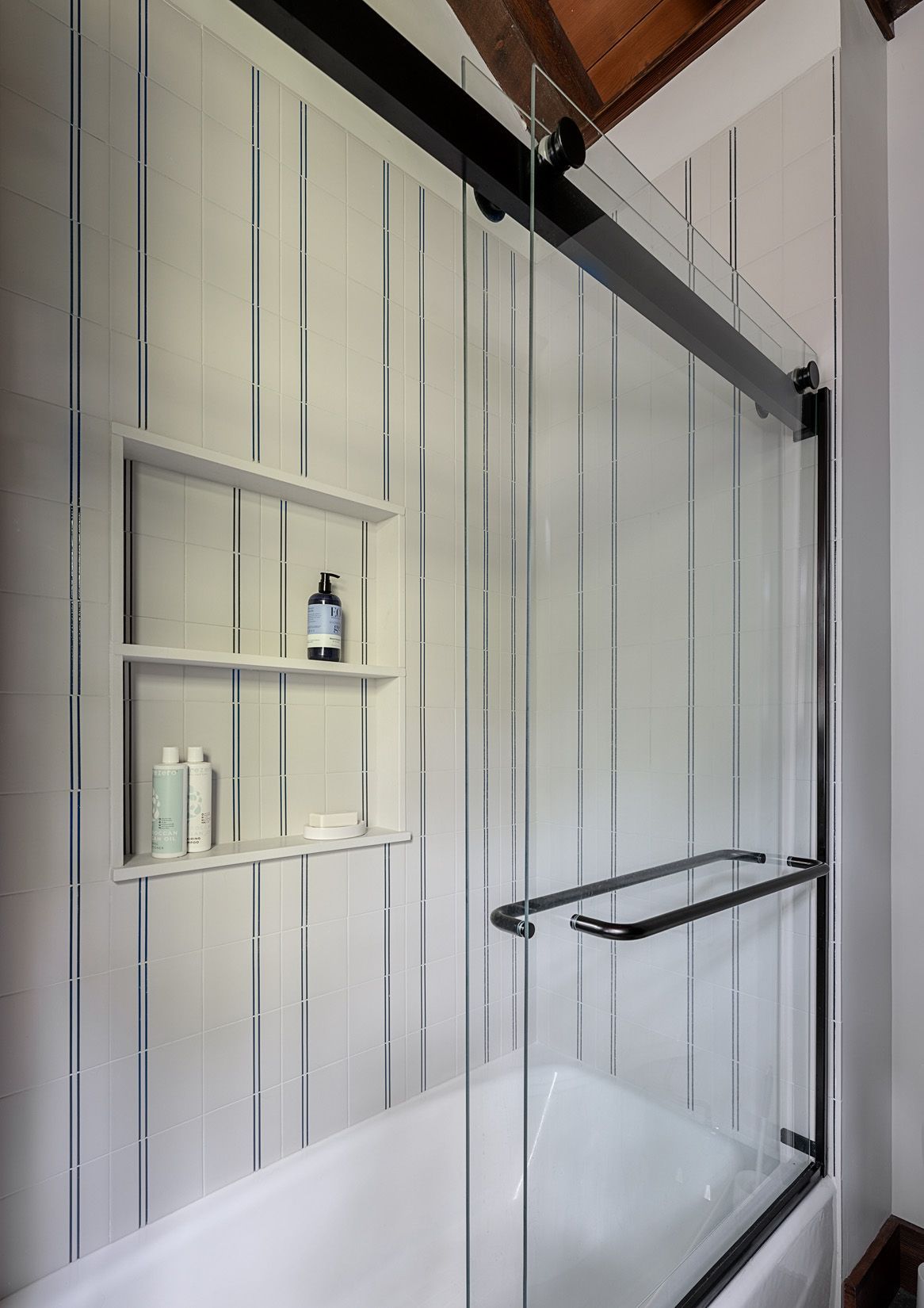 A modern shower with white vertical-tiled walls, a built-in recessed shelf with toiletries, and a sliding glass door.