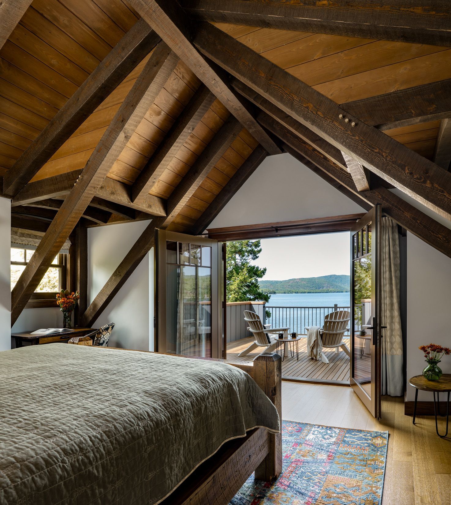 A bedroom with vaulted wooden beams and a bed overlooking a balcony with chairs and a lake view.