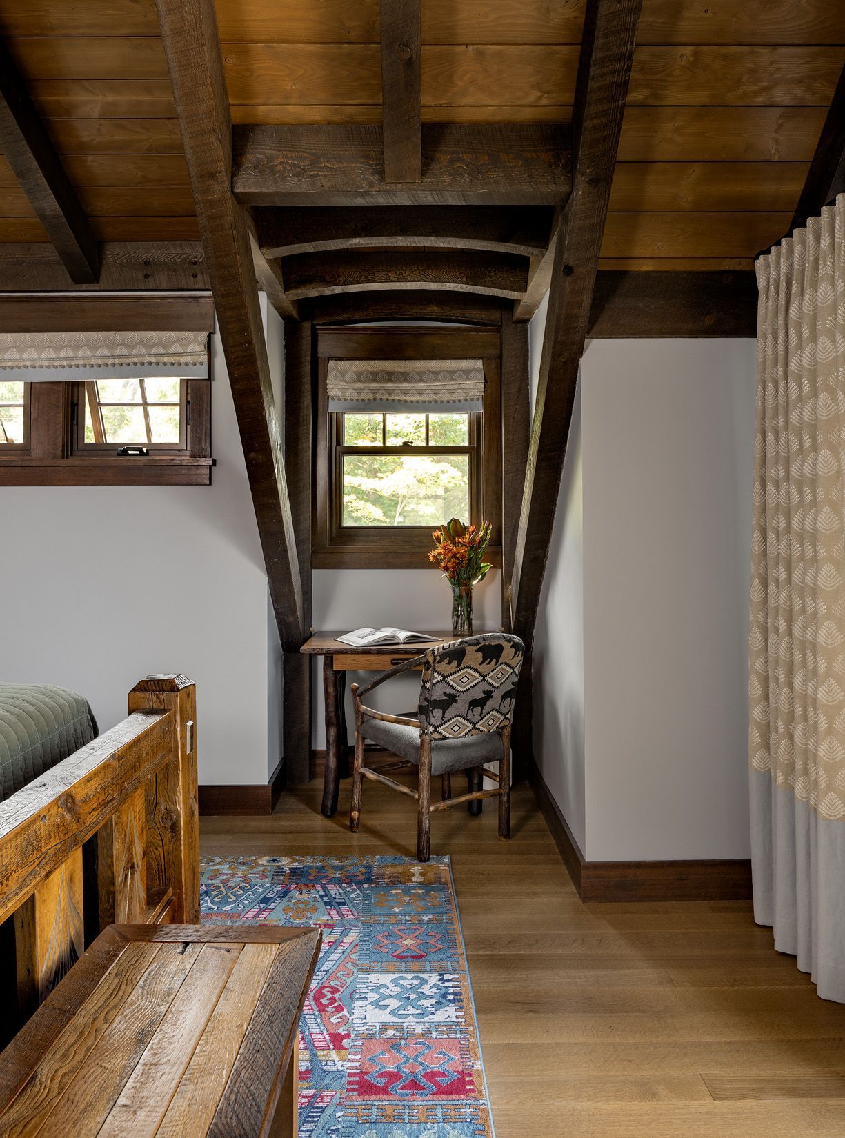 A cozy nook with a wooden desk and patterned chair under a window, framed by dark wood beams in a rustic room.