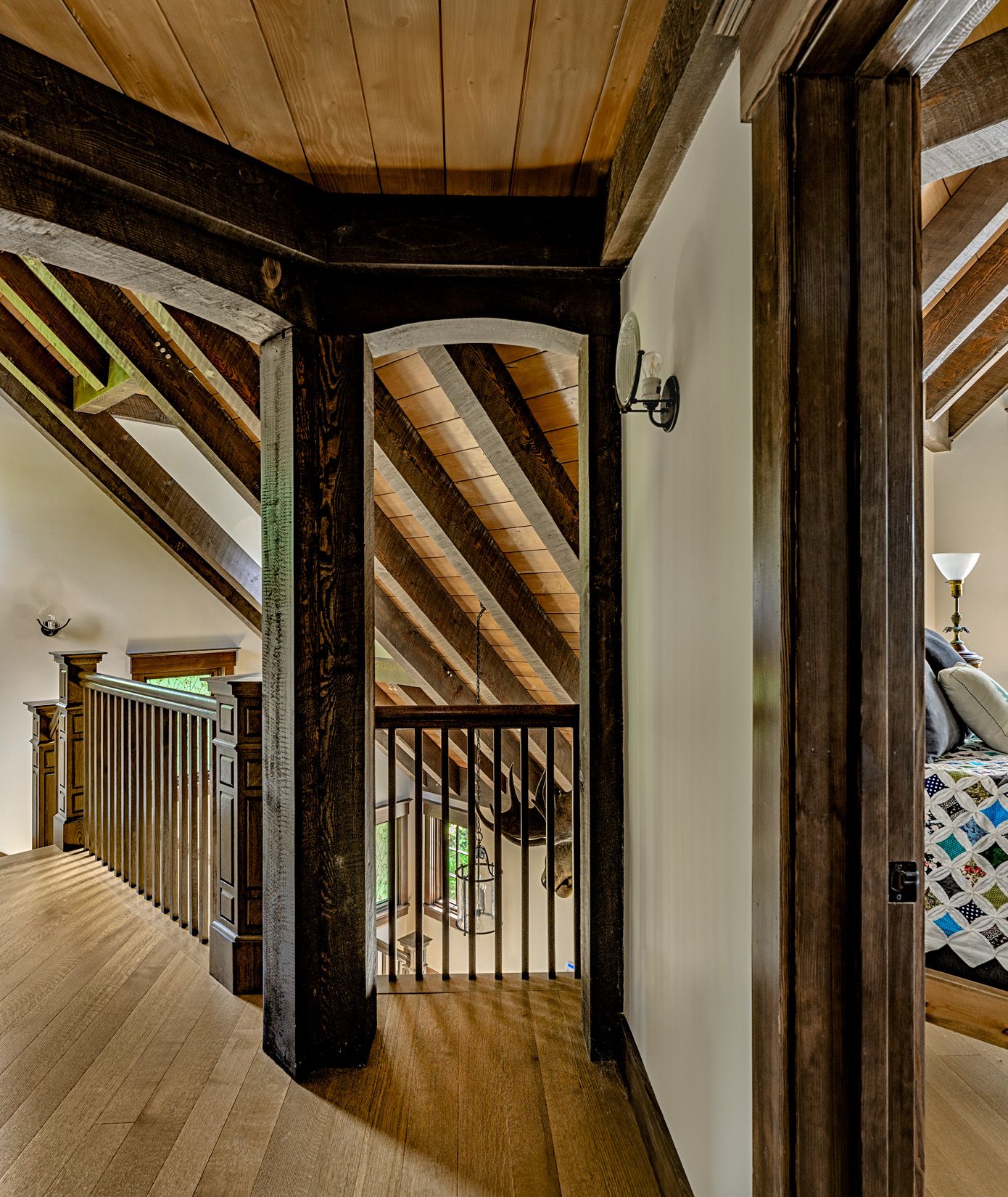 An indoor loft landing with wooden floors, dark timber beams, sloped ceilings, and wooden railings overlooking a room.