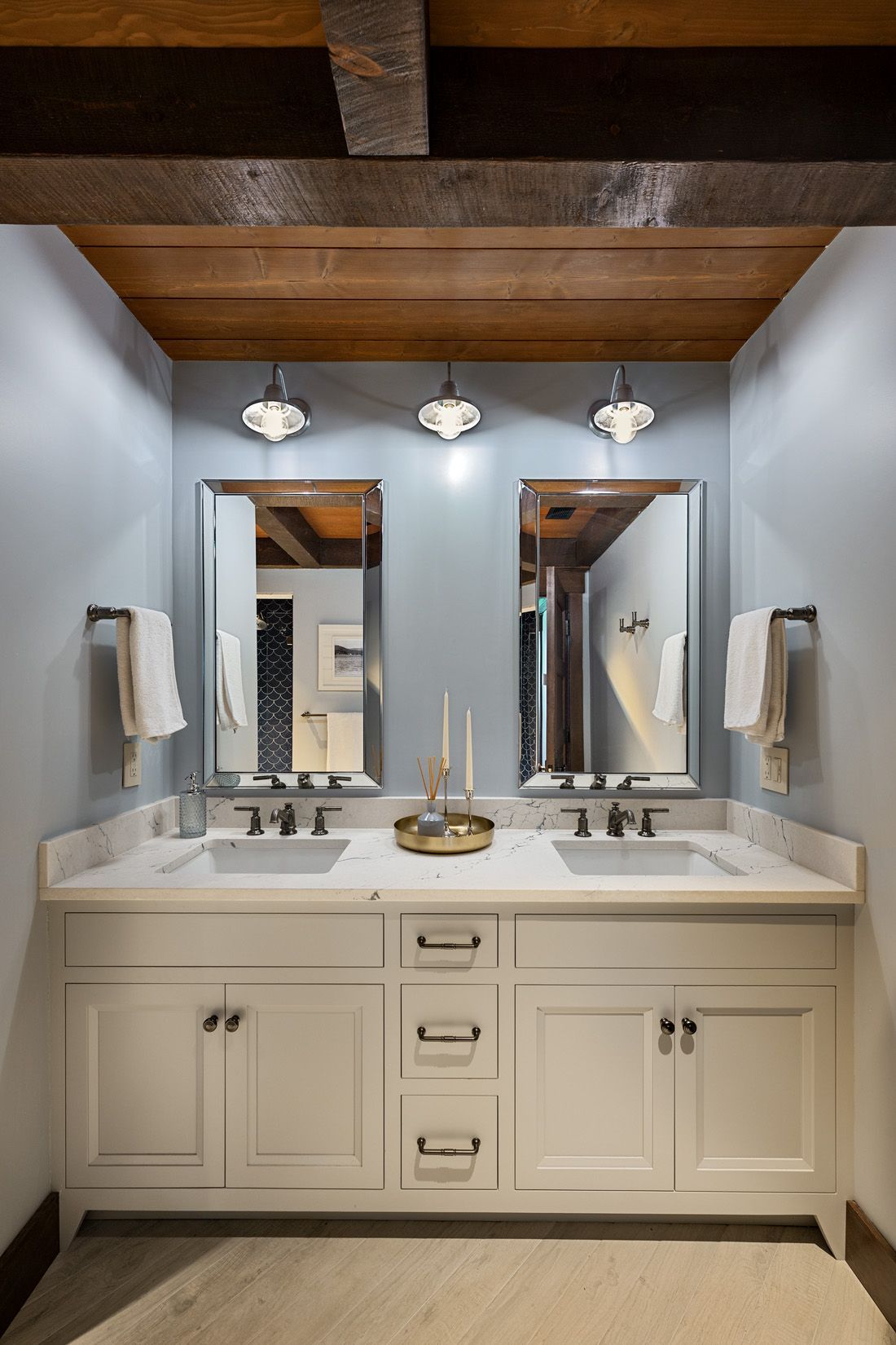 A double-sink bathroom vanity with white cabinets, light blue walls, two framed mirrors, and a wooden beam ceiling.