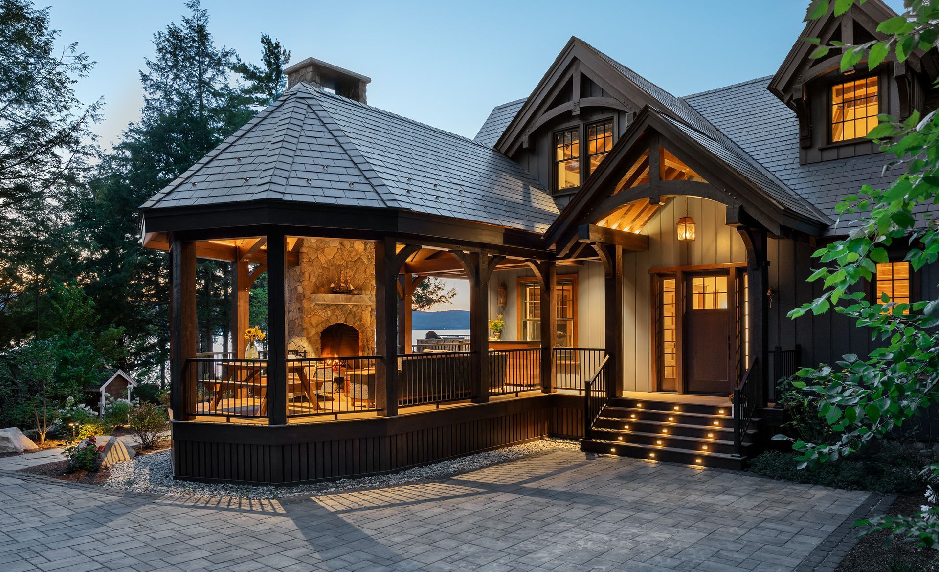 A cozy, rustic lake house with a large stone fireplace on an illuminated porch, stone patio, and trees at dusk.