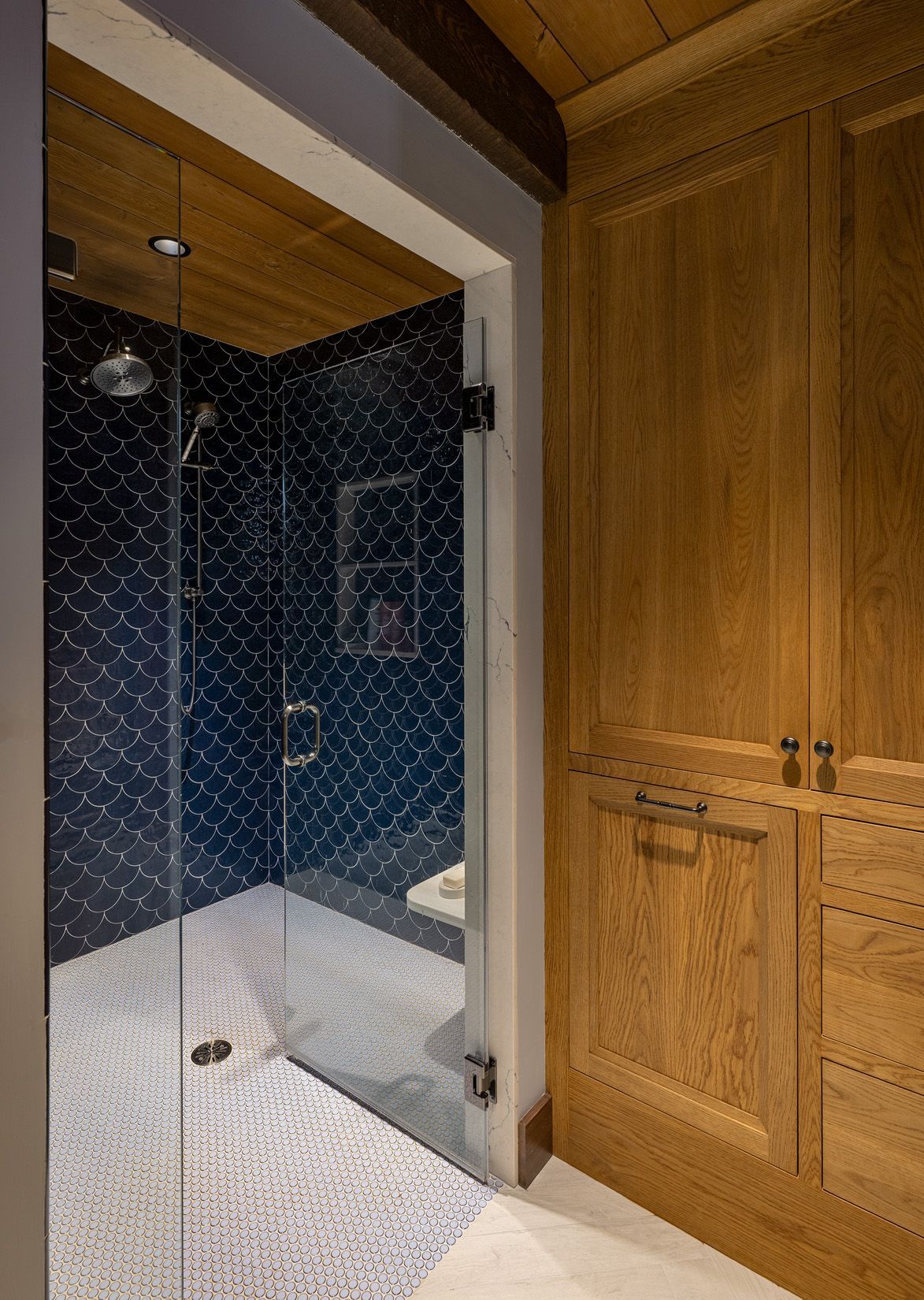 A modern shower with dark blue patterned tile, a glass door, and wooden cabinetry beside it in a bathroom.