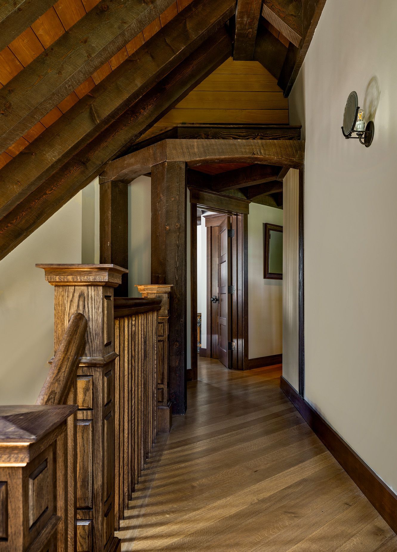 A narrow hallway with a wooden vaulted ceiling, decorative railing, and hardwood floors leading to a dark wood door.