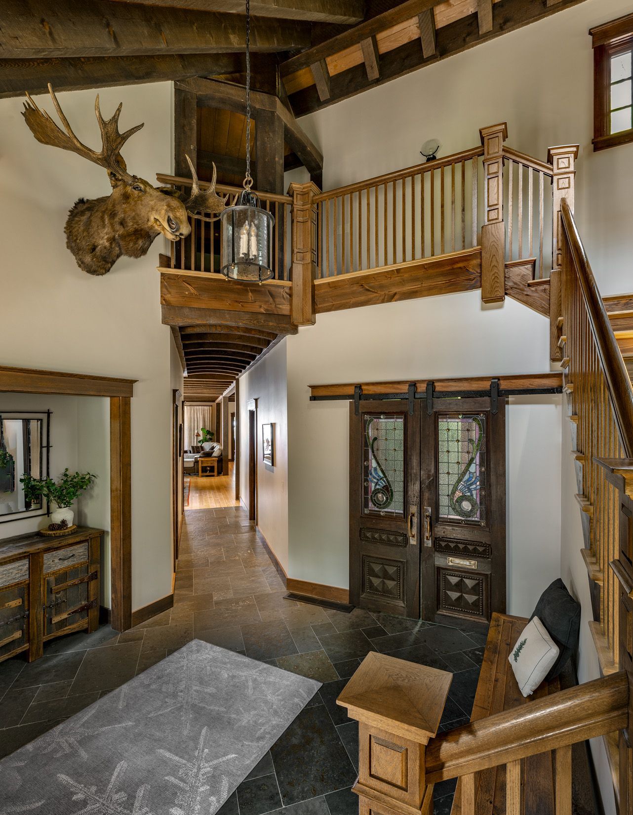 A rustic, wood-trimmed foyer with a moose mount on the wall, a stone floor, and a wooden staircase leading to a loft.