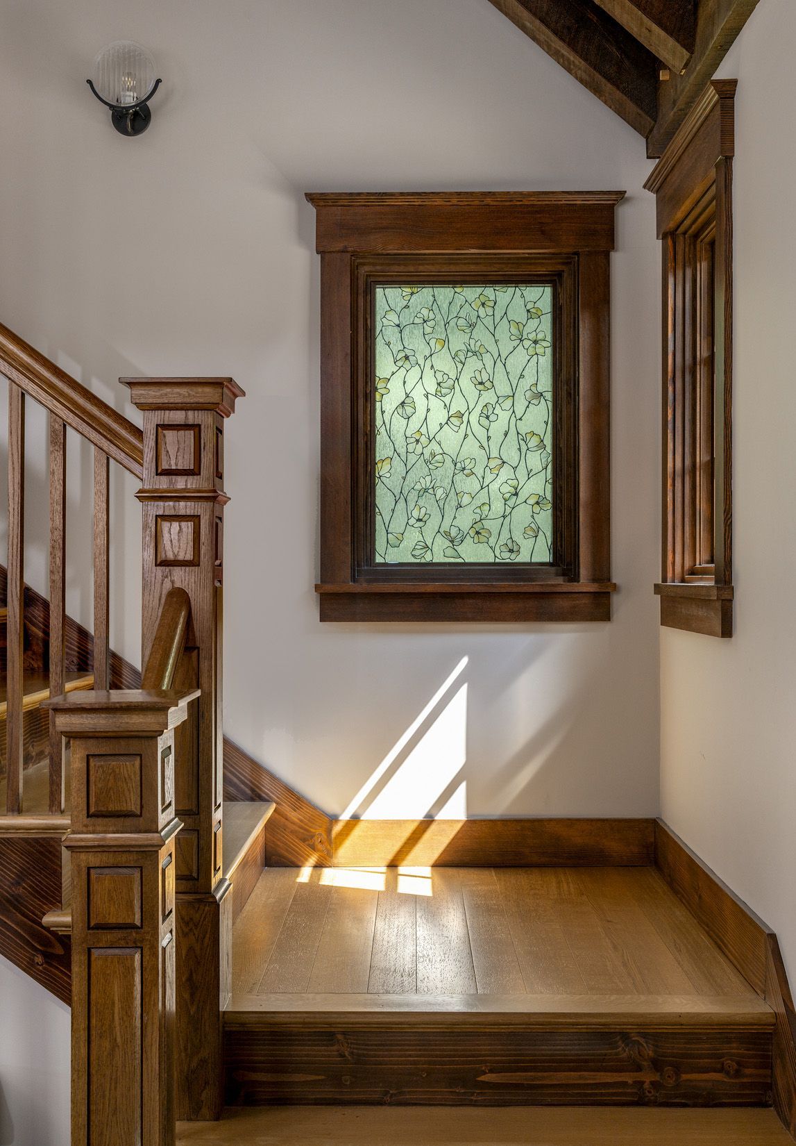 Staircase landing with wooden railing, trim, and stairs illuminated by sunlight through a patterned window.