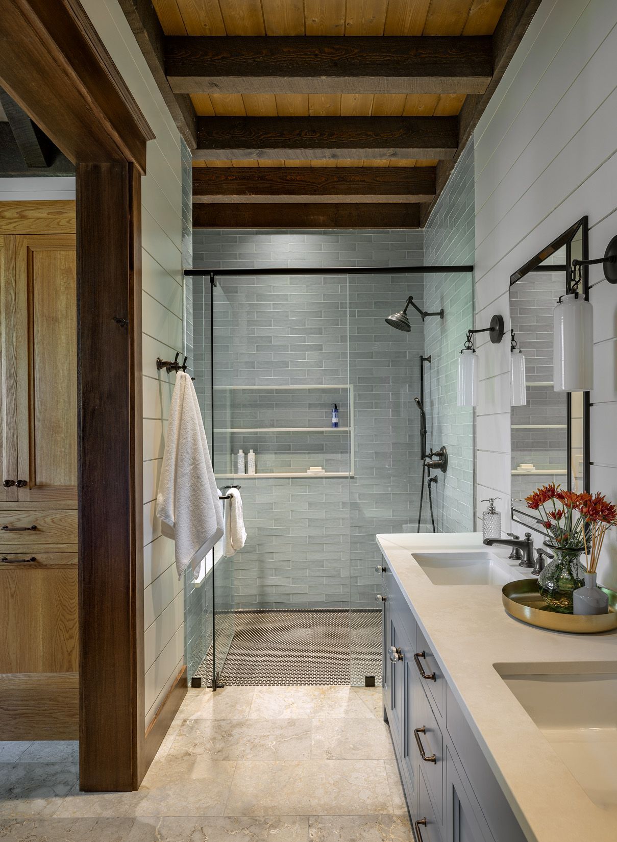 A modern bathroom featuring blue-grey tiled walk-in shower, double vanity, light stone floors, and exposed wood ceiling.