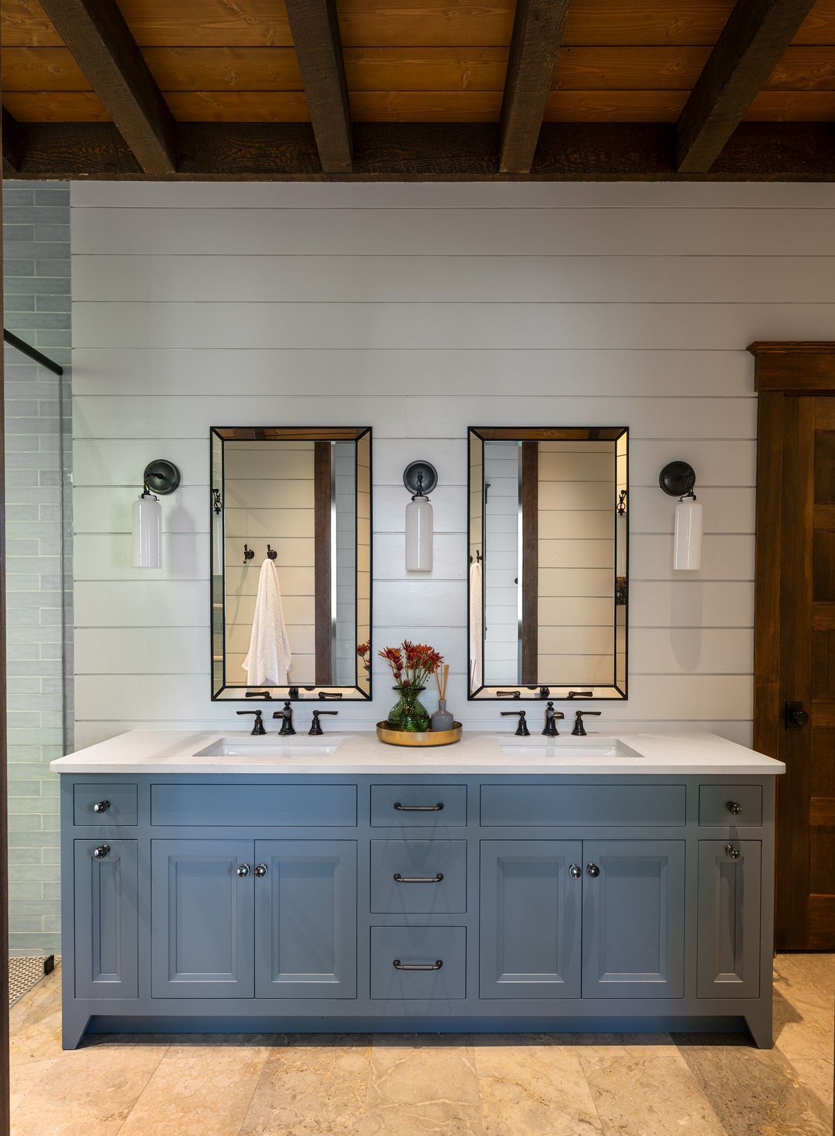 Double-sink bathroom vanity with grey cabinets, white shiplap walls, twin mirrors, and wall sconces under wood ceiling beams.