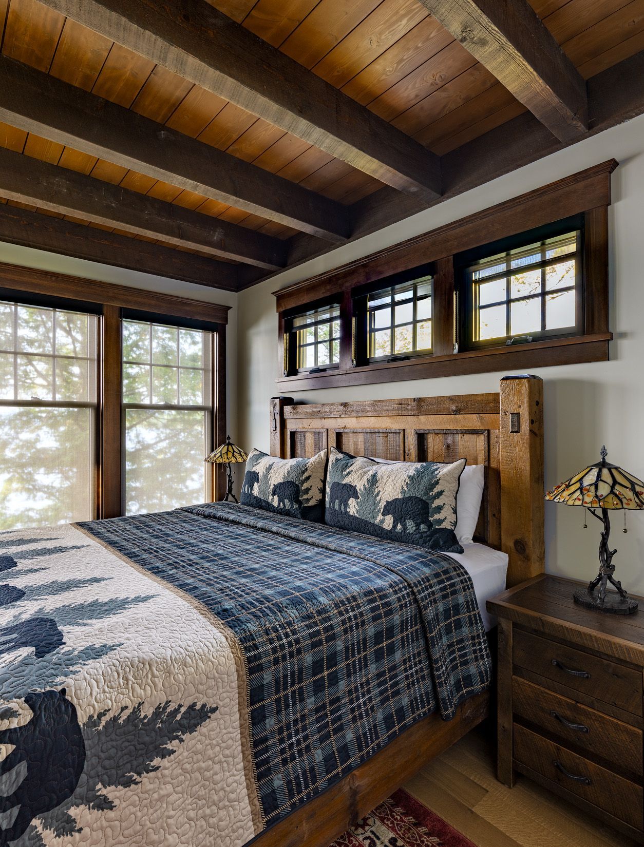 Rustic bedroom with a wooden bed frame, bear-patterned bedding, exposed ceiling beams, and small windows above the head.