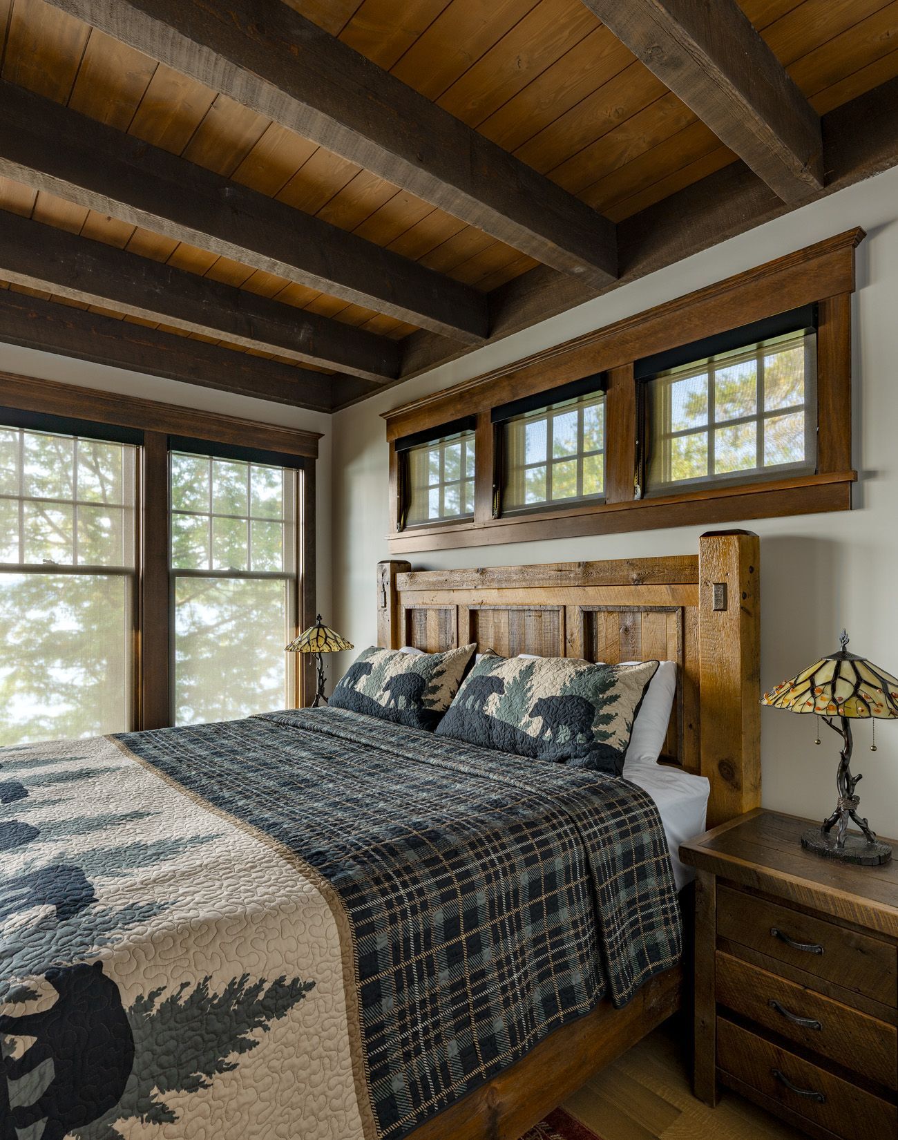 A rustic bedroom featuring a wooden bed frame, a patterned bear quilt, and a stained-glass lamp beside a windowed wall.