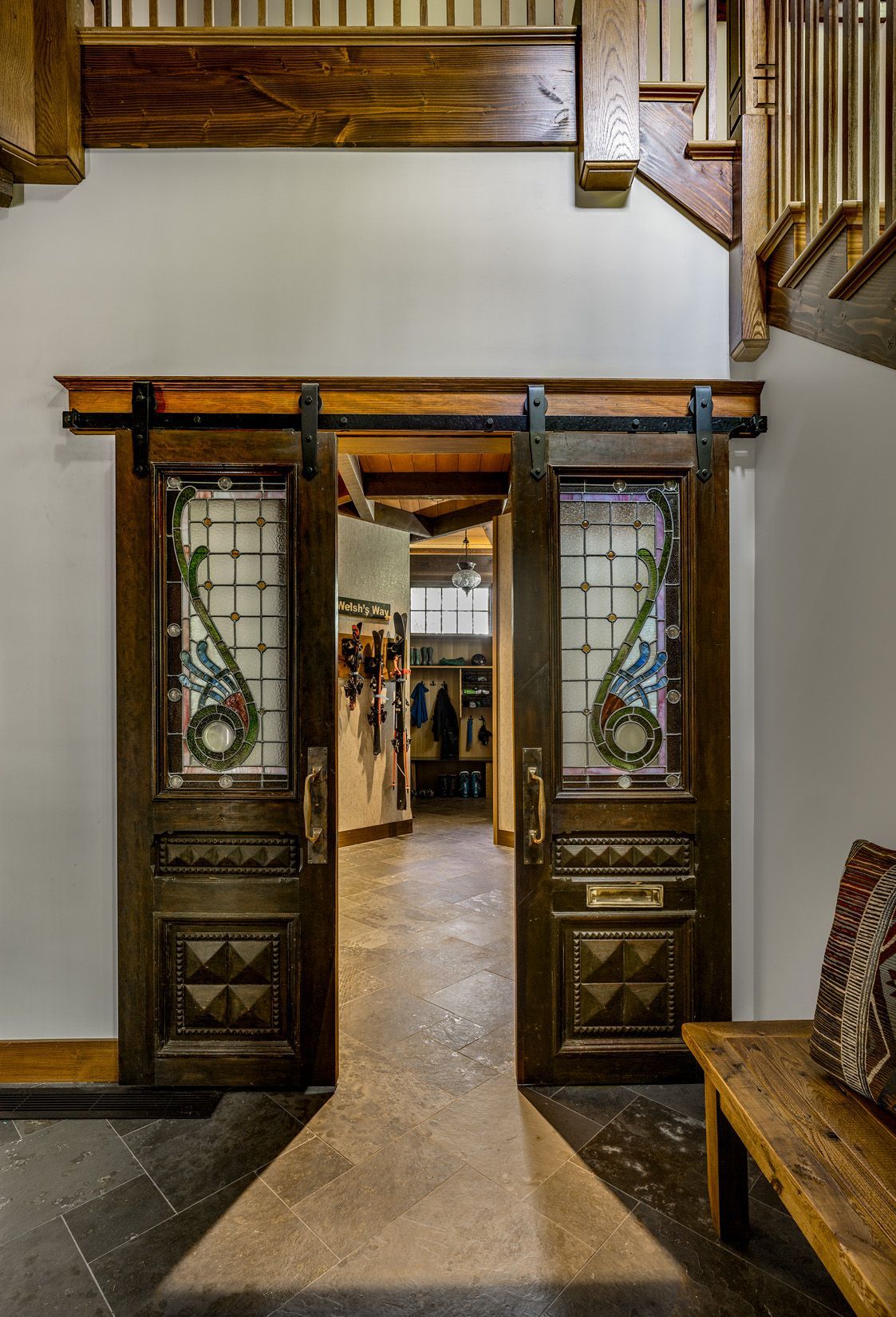 Double wooden barn-style doors with stained glass panels open to a tiled hallway under an indoor balcony.