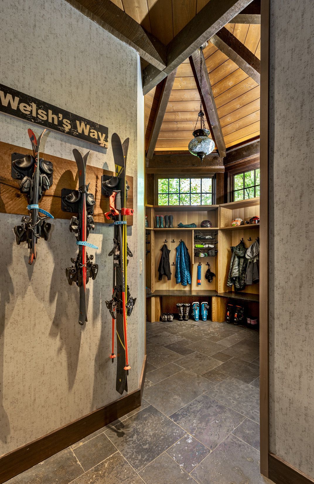 A ski mudroom with a stone floor and wood-paneled walls features racks of skis and cubbies with hanging coats and boots.