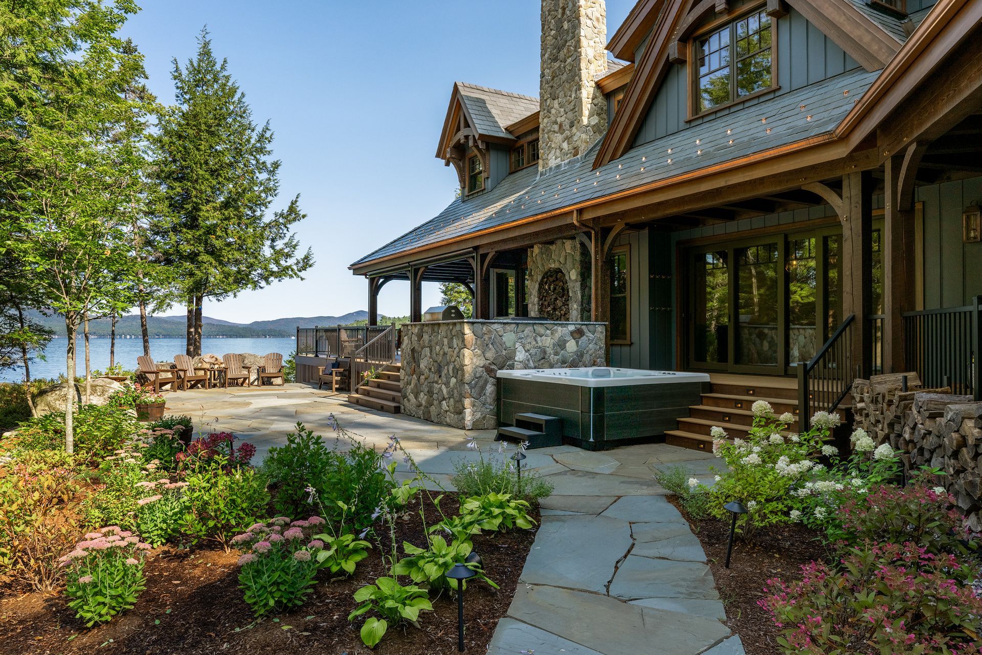 A rustic stone and wood home featuring a backyard patio, a hot tub, and lush gardens, overlooking a lake.