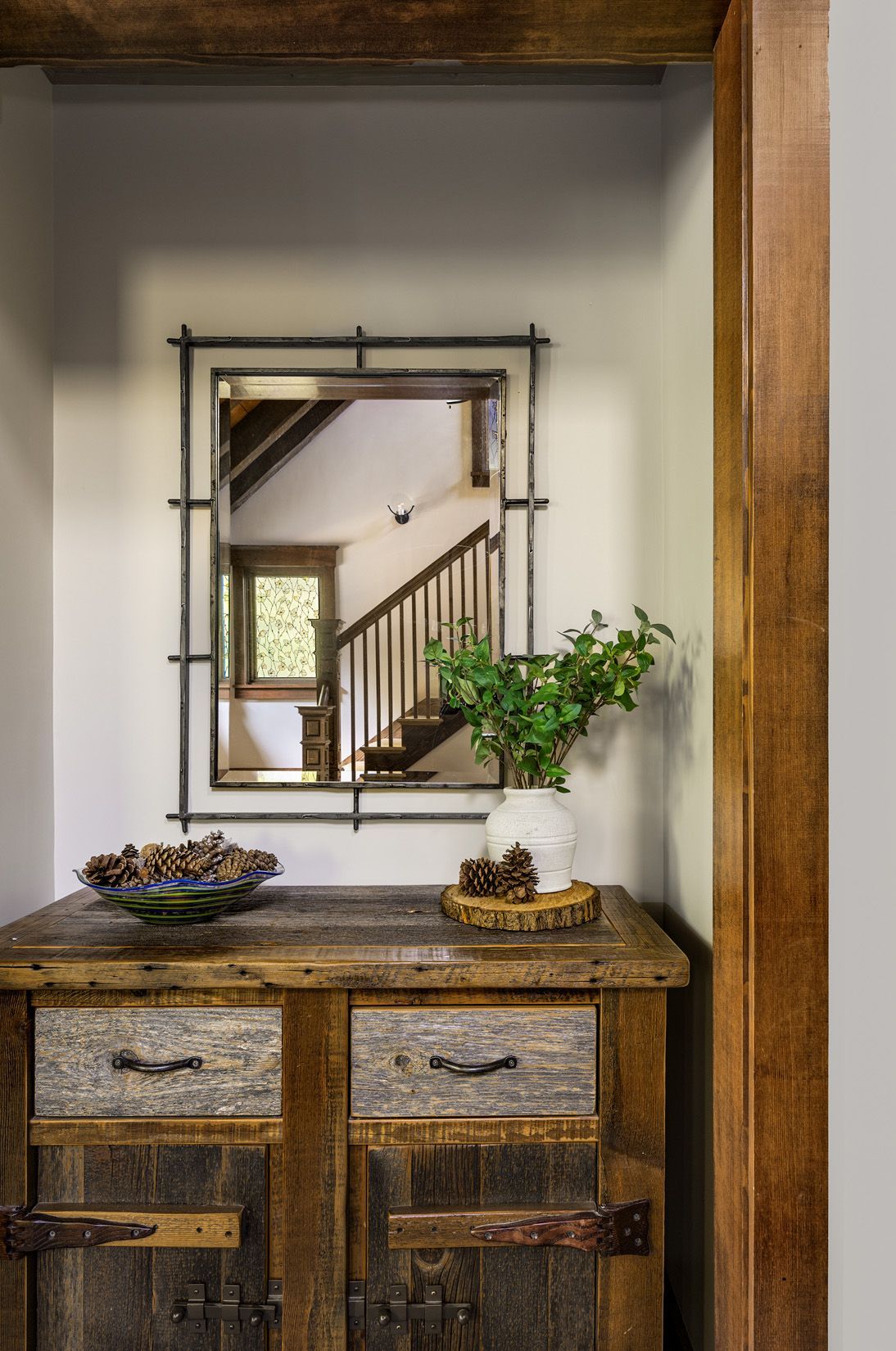 A rustic wooden cabinet with two drawers and doors sits below a rectangular, metal-framed mirror reflecting a staircase.