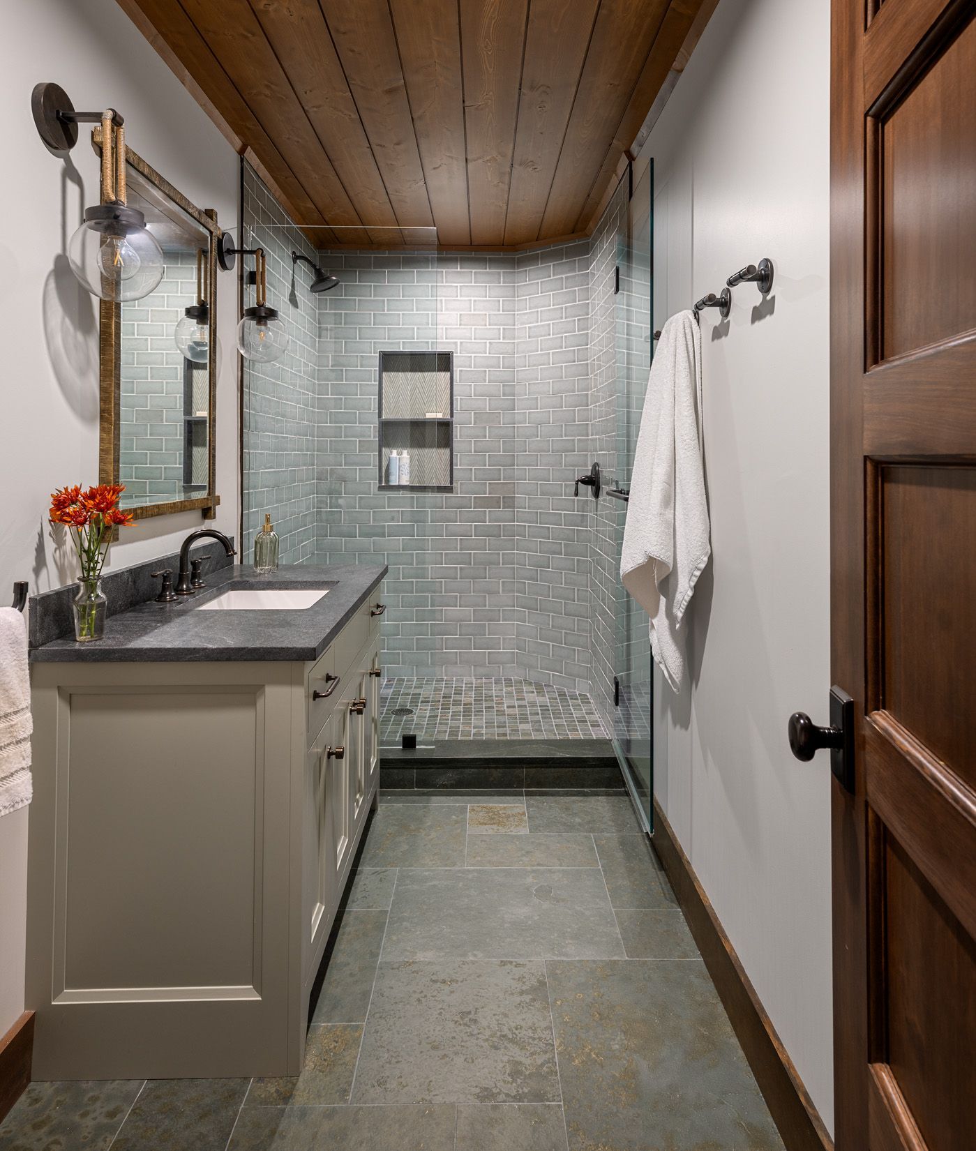 A bathroom with a gray vanity, stone tile floor, wood-paneled ceiling, and a glass-enclosed shower with subway tiles.
